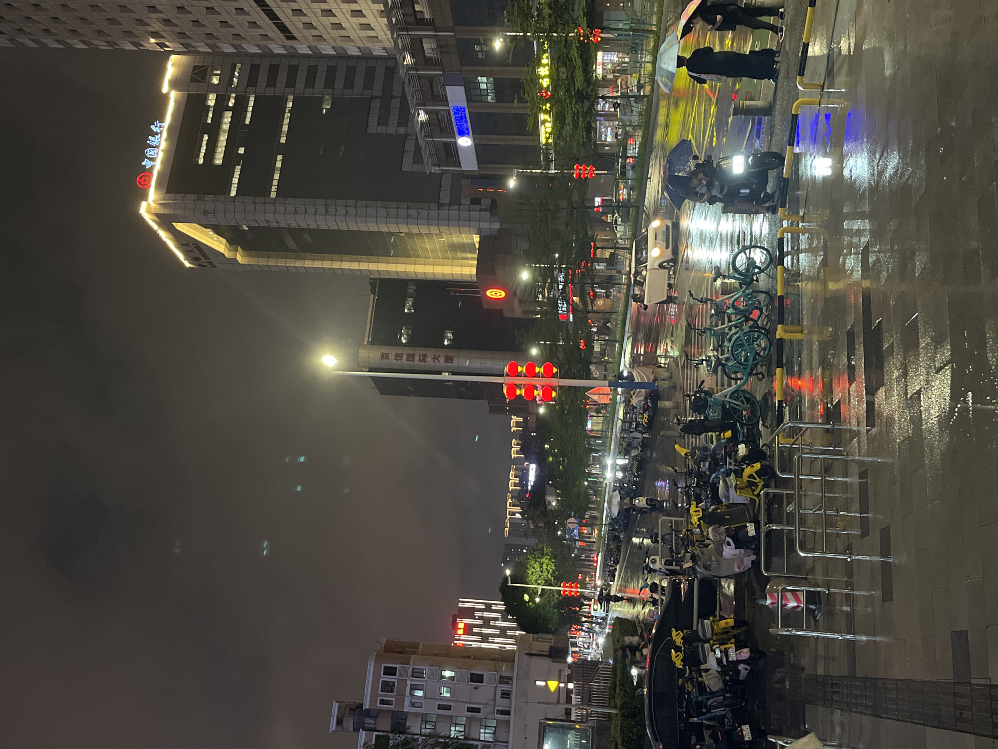 This image depicts a street scene at night in the Nanhai District of China, likely during or after rainfall, as indicated by the wet pavement and reflections. Tall buildings with illuminated windows and signage dominate the background. One prominent building features a neon sign with Chinese characters and a circular red logo at its peak. A traffic light with red signals is visible, suggesting vehicle traffic. The street is lined with numerous parked bicycles and scooters, many of which are yellow. Several individuals are present, some holding umbrellas, and a few are riding or standing near scooters. A white car is visible in the street. The overall atmosphere is urban and commercial, with the wet conditions creating a luminous quality on the ground.