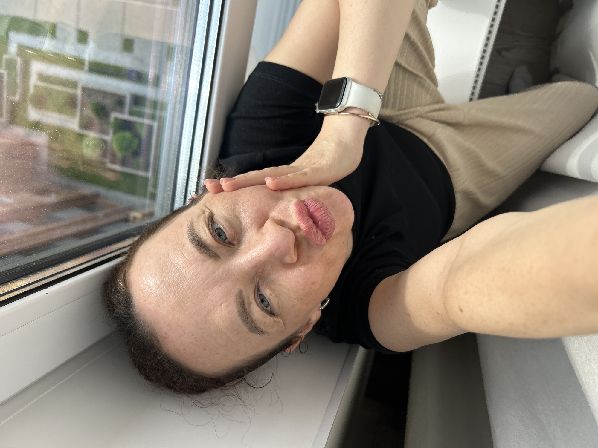 In Yekaterinburg, Russia, a woman is taking a selfie by a window. She is wearing a black t-shirt and tan pants, and a white smartwatch on her left wrist. She is leaning her head against her hand and making a kissing face, with her lips puckered. The window behind her offers a view of a courtyard with geometric landscaping. The overall scene is casual and personal.