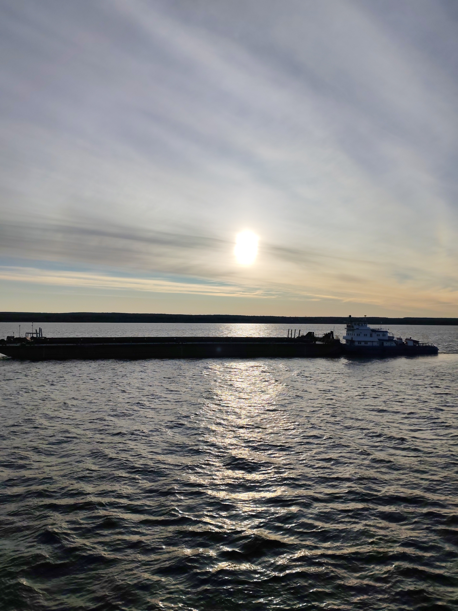 A long, flat barge is on the water under a sky where the sun is visible. At the end of the barge is a tugboat. The location is Shchetinskoe, Russia. The water is rippled, and the sun's reflection creates a shimmering effect on its surface. The horizon shows a dark line of land. The sky has light clouds with a hint of yellow from the sun.