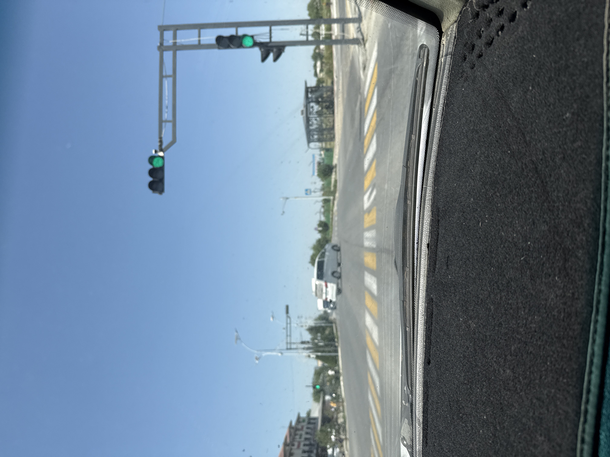 The image shows a view from inside a vehicle, looking out at a street scene. The dominant colors are blue from the sky and grey from the road and traffic signals.
The main subjects are traffic lights, a road with pedestrian crossings, and a few vehicles. There are two sets of traffic lights visible. The one on the left has a green light illuminated, and the one further down the road also appears to have a green light. Several vehicles are present, including a white van or bus and possibly other cars, though they are out of focus. The road has clear yellow and white striped pedestrian crossings. In the background, there are buildings and trees, suggesting an urban or semi-urban environment. A street lamp is also visible.
The image appears to capture a moment during daytime, with a clear blue sky and bright sunlight, indicating fair weather. The presence of traffic lights and vehicles suggests that this is a street or intersection where traffic is regulated. The perspective from inside the car indicates that the vehicle is either stopped at a red light or moving slowly through the intersection.
There is no visible text in the image. The location context of Pakhtakor, Uzbekistan, provides additional information about the setting.
