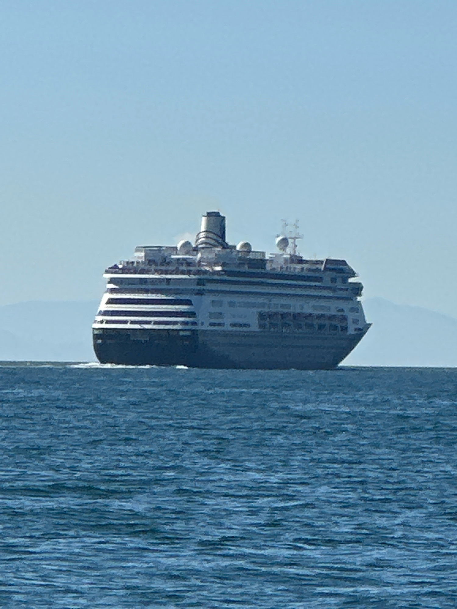 A large cruise ship is sailing on a body of water. The ship is primarily white and gray, with multiple decks and several windows visible. It has a distinctive smokestack with a ribbed design. The ship is moving from left to right, creating a wake behind it. In the background, there are hazy mountains and a clear blue sky. The water in the foreground is dark blue with visible ripples. The image appears to be taken from a distance, likely from another boat or the shore.
This scene is likely located in the waters off the coast of West Vancouver, Canada, given the mountainous backdrop characteristic of the region.