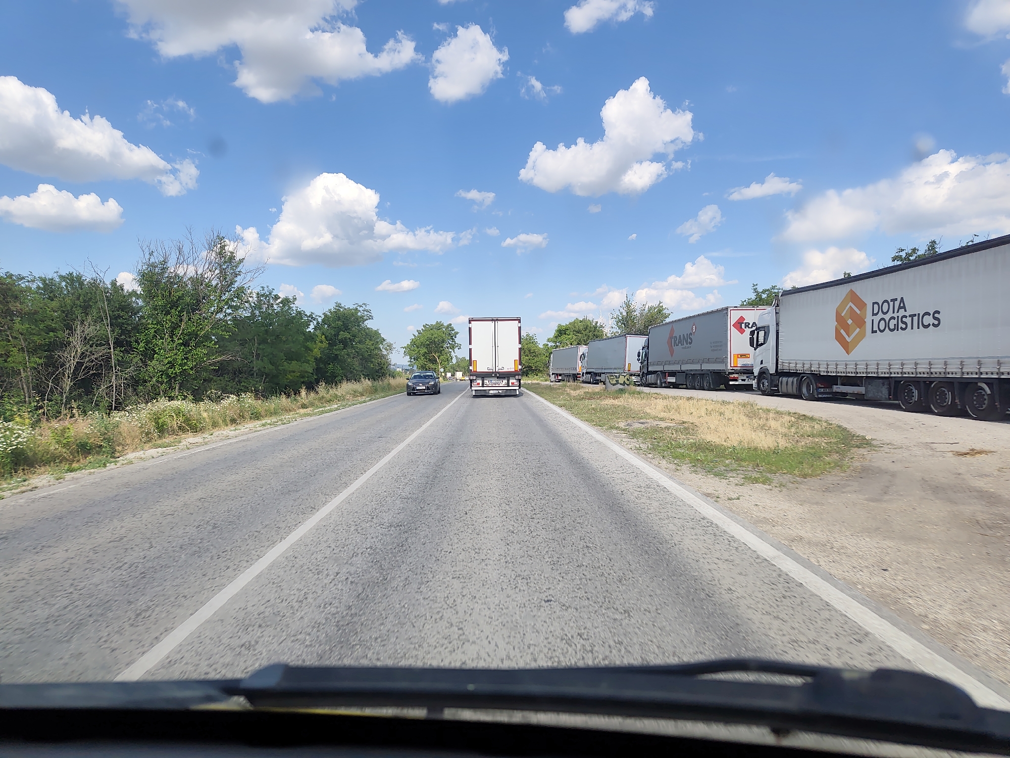 A forward-facing, wide-angle, eye-level, full shot captures a sunny day on a two-lane asphalt road with a white line dividing the lanes. The road is flanked by green trees and dry grass. On the right side of the road, a convoy of white semi-trailer trucks are parked, with "TRANS" and "DOTA LOGISTICS" logos visible on some. A black sedan is traveling in the left lane ahead. The sky is a vibrant blue with scattered white clouds. The foreground shows the edge of a vehicle's dashboard. The image appears to be taken from a moving vehicle, suggesting a view of a roadside in the vicinity of Obnova, Bulgaria, where such logistics operations might occur.