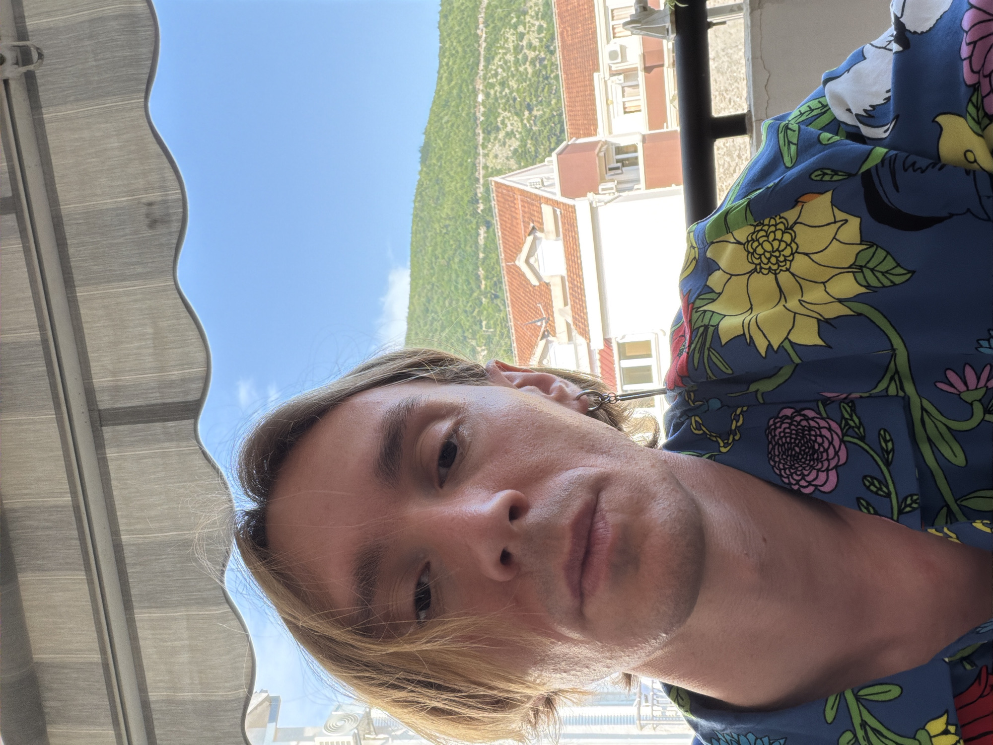 In Budva, Montenegro, a close-up selfie shows a person with blonde hair and an earring. They are wearing a blue shirt with a colorful floral pattern, featuring yellow, pink, and green flowers. The background includes buildings with red tile roofs, lush green hills, and a bright blue sky with a few clouds. An awning is visible above the subject.