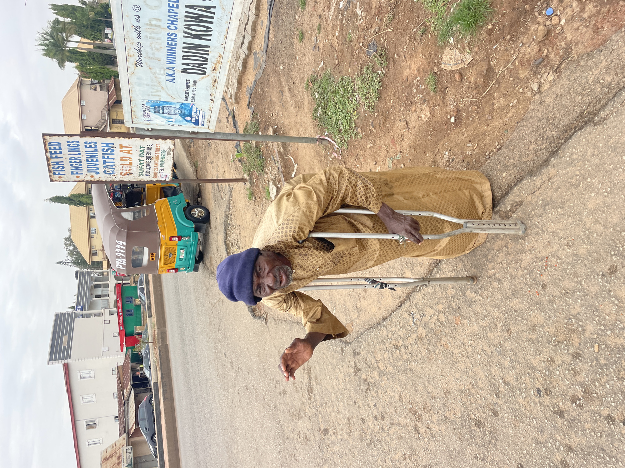 The image depicts an elderly man with a blue knit cap and wearing a patterned tan robe standing near a road using crutches. The man appears to be smiling at the camera. Beside him is a sign for "FISH FEED, FINGER LINGS, JUVENILES CATFISH SOLD AT SUFAT DAT." A sign above this advertises A.K.A Winners Chapel in Dadin Kowa with Sunday service hours.
Several vehicles including green and yellow auto rickshaws (tuk-tuks) and cars are parked or traveling along the road. The setting appears to be Jos, Nigeria, based on contextual clues from the text visible on the signage.