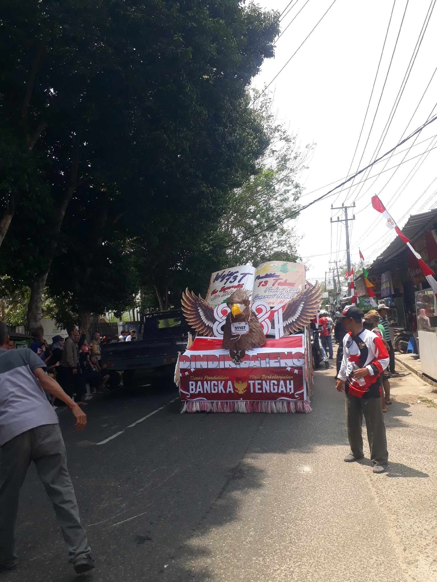 In Pangkalpinang, Indonesia, a parade is underway. The foreground features a person walking on the right side of the image, wearing a white and red patterned shirt and dark pants. They are holding a water bottle. To the left, another person is walking away from the camera, wearing a grey shirt and grey pants. In the background, a decorated float is moving down the street. The float has large eagle wings and a depiction of an eagle with the number "51107" on its chest. Text on the float reads "DINAS PENDIDIKAN" and "BANGKA TENGAH". Several people are visible on and around the float, as well as spectators lining the street. Indonesian flags are displayed on poles along the road. The scene is set on a paved street with trees and buildings visible on either side, under a bright, overcast sky.