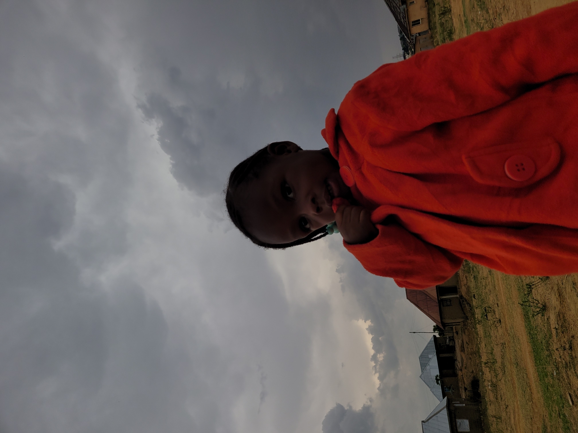 A girl stands in an outdoor scene. She wears an orange coat. Behind her, the sky is overcast with grey clouds. Some buildings are visible in the background, indicating a setting in Barakin Akawo, Nigeria. The ground appears to be grassy or dirt.