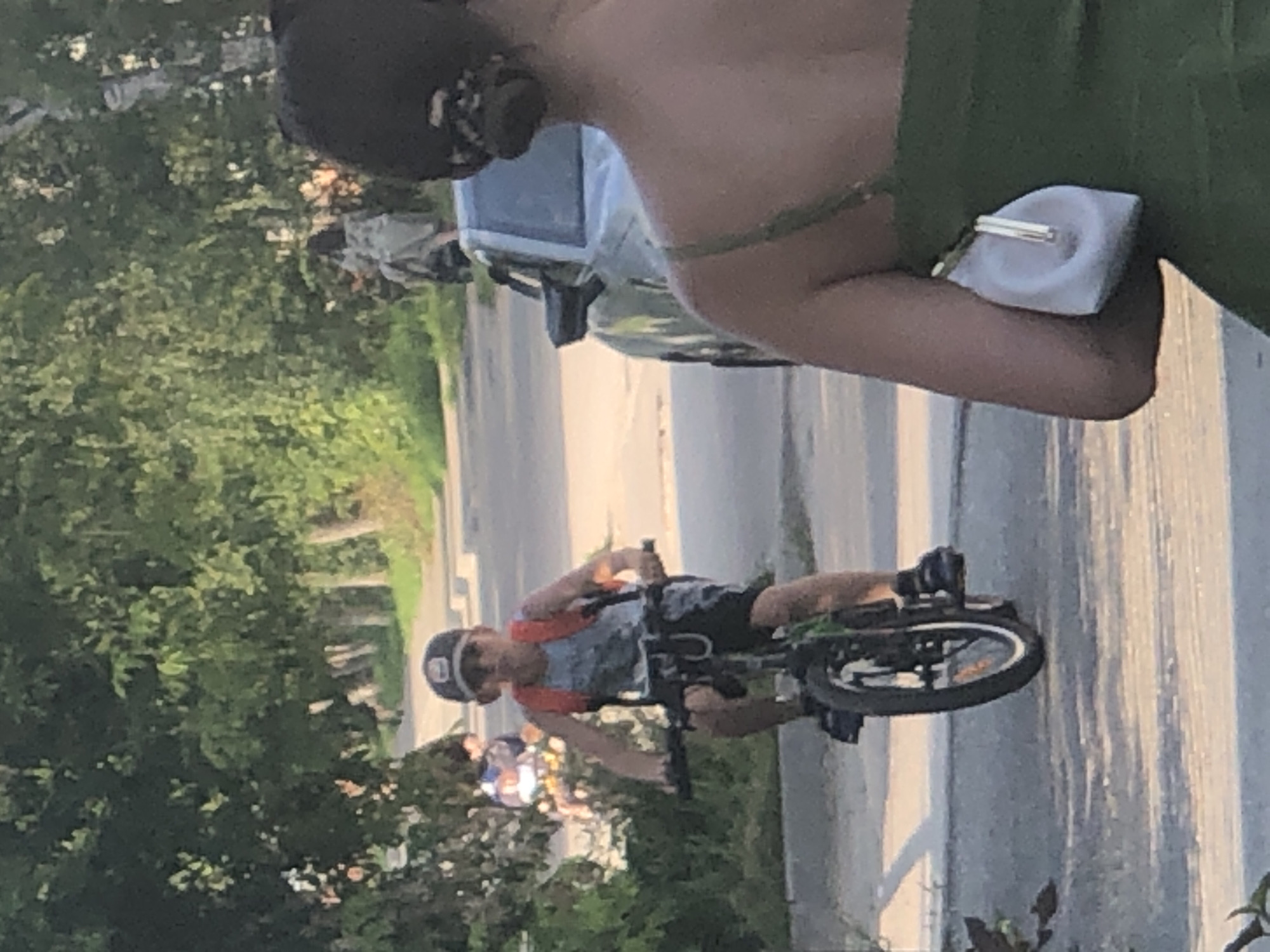The media file shows a young boy riding a bicycle. He is wearing a helmet and a t-shirt with a colorful pattern and orange straps. The boy is positioned in the foreground of the image, with trees and a road visible in the background. To the left of the boy, there is another child on a bicycle, also wearing a helmet. Further in the background, a car is parked, and it appears to be a light-colored sedan. The scene is outdoors, likely during the daytime, as indicated by the natural lighting and the shadows cast. The overall impression is of a casual, recreational activity taking place in a residential or park-like setting. The image is taken from a slightly elevated angle, looking down at the boy.