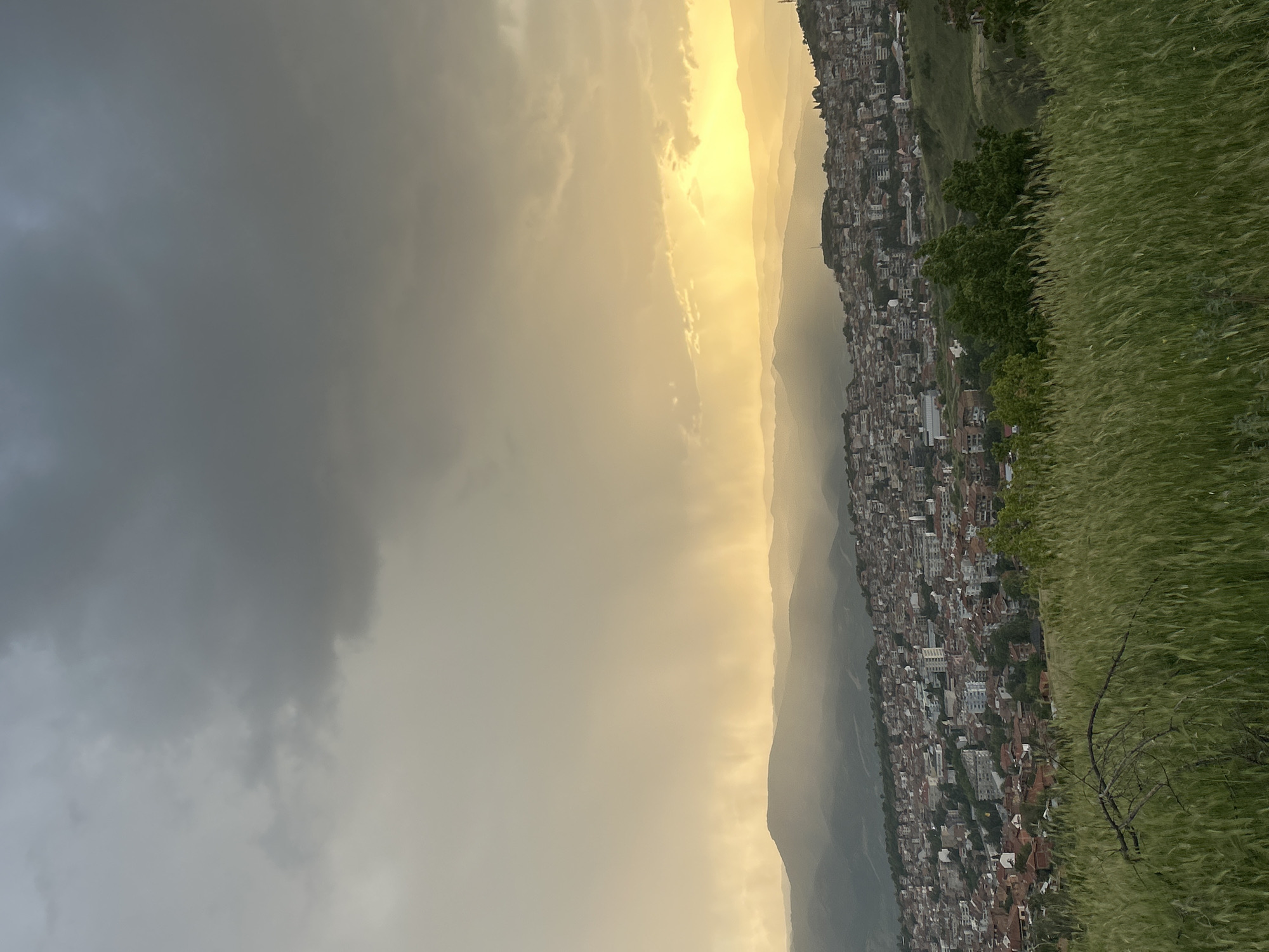 Captured from an elevated perspective, the landscape features the city of Polenitsa, Bulgaria. The city sprawls over mountainous terrain, characterized by dense residential buildings with reddish rooftops. Transitioning from the built environment is verdant green grass. The sky above is divided between a heavy, overcast gray area and a luminous, golden-hued region where the sun appears to pierce through the atmospheric haze.