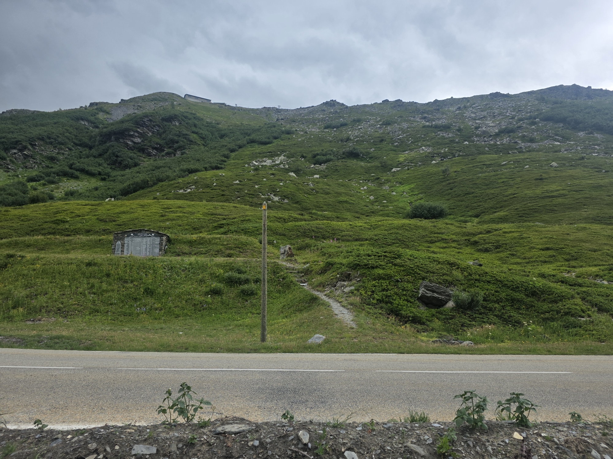 The image shows a mountainous landscape with a road in the foreground. The mountain slopes are covered with green vegetation and rocks. A small, rustic building with a memorial plaque is visible on the slope. A wooden pole with a yellow marker stands next to a path leading up the mountain. The sky is overcast. The image appears to be taken in the region of Séez, France.