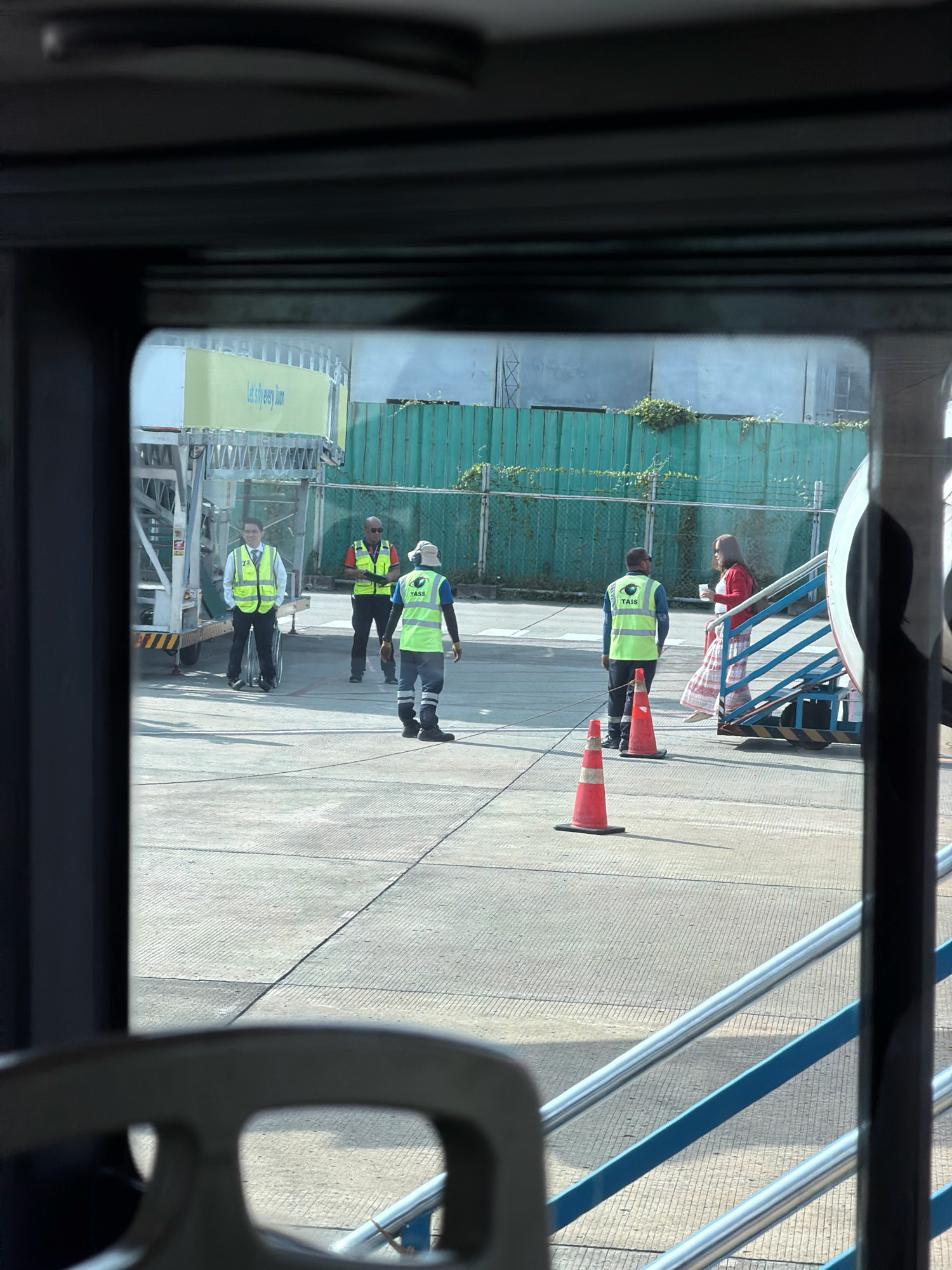 This is a photograph taken from inside a vehicle, looking out through a window at an airport tarmac. In the foreground, the dark interior of the vehicle is visible, including a gray, worn armrest. The main scene shows several individuals on the tarmac. To the left, a person in a yellow vest and dark pants stands near a piece of airport equipment. Further to the right, three people are visible. Two are wearing high-visibility yellow vests and dark pants. One of these individuals is speaking with a person wearing a red jacket and a patterned skirt, who is standing on an airstair. Orange traffic cones are placed on the tarmac. In the background, a green fence with overgrown vegetation runs along the edge of the tarmac, and buildings are visible beyond it. A large yellow banner with the text "Let's fly every Boar" is attached to airport equipment. The lighting suggests it is daytime. The location is specified as Nabas, Philippines.