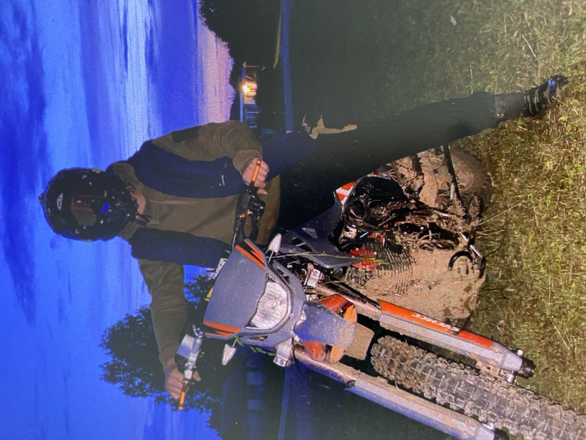 The image captures a person standing with a heavily mud-covered dirt bike in an outdoor setting during the evening or night. The individual, appearing male, wears a dark full-face helmet, an olive green or brown long-sleeved jacket over a dark blue garment, dark trousers, and dark boots. He is positioned on or beside an orange and grey dirt bike, holding the handlebars.
The motorcycle is thoroughly caked in thick mud across its frame, engine, wheels, and suspension components, particularly around the front wheel and engine block. Its knobby tires are also laden with mud. The front headlight assembly is visible, showing parts of the grey and orange fairing.
The background is dominated by a dark blue sky, indicative of dusk or nighttime. Trees are visible on the horizon, and faint artificial lights can be seen in the distance, suggesting a nearby road or structures. The ground immediately surrounding the bike is a mix of grass and deep mud. Faint white lettering, possibly reading "RANCO", is discernible on the right sleeve of the person's jacket. The person's face is obscured by the helmet, leaving their expression unreadable. The scene portrays a moment during or after an off-road adventure through muddy terrain.