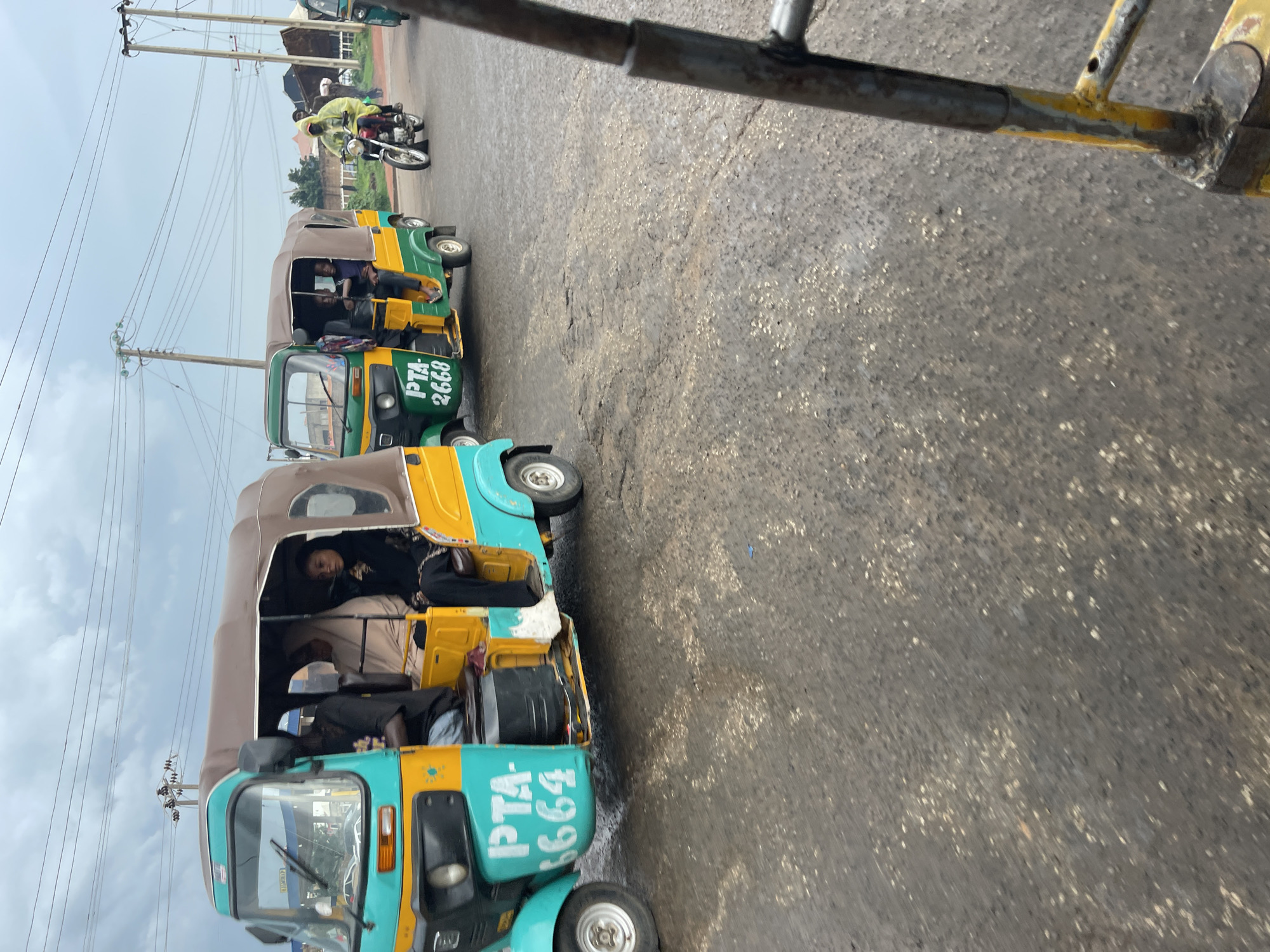 The image shows a street scene in Jos, Nigeria, with several auto-rickshaws, also known as Keke NAPEPs, and a motorcycle. The Keke NAPEPs are predominantly painted in green and yellow or turquoise and yellow. Passengers are visible inside the Keke NAPEPs, some looking out at the camera. One Keke NAPEP has the text "PTA-2668" painted on its side, and another has "PTA-2664". A motorcycle with a rider wearing a yellow rain jacket is also present. The road surface appears wet and worn, suggesting recent rain or damp conditions. The sky is overcast with visible utility poles and power lines in the background, indicating an urban or semi-urban setting. The overall mood appears to be that of daily commute or transportation. The time of day is difficult to ascertain precisely due to the overcast sky, but it seems to be daytime.