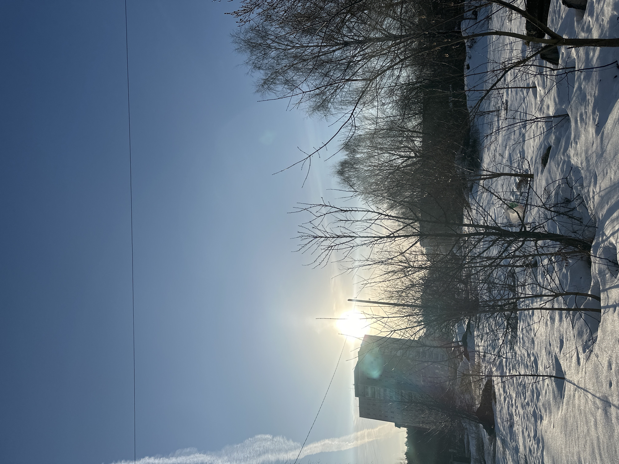 The image depicts a winter scene in what is likely Chaikovsky, Russia, given the request context. The photograph is oriented vertically. The scene features a snow-covered landscape with bare trees silhouetted against a bright, sunlit sky. A multi-story building is visible in the foreground, its facade appearing somewhat aged and indistinct due to the light. The sun is positioned behind the building, creating a lens flare effect. A prominent power line stretches across the left side of the sky. In the lower-left, contrails are visible. The ground is covered in deep snow, with shadows cast by trees and possibly other structures. The overall impression is one of a cold, sunny winter day in an urban environment. No people are visible in the scene.