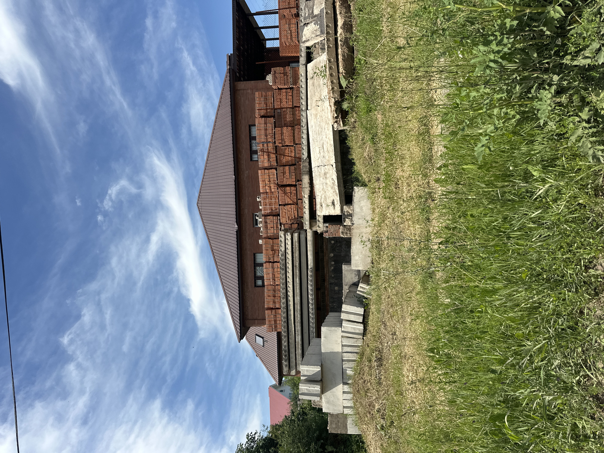 A brick house with a brown metal roof is under construction in Sterlitamak, Russia. In front of the house, there are stacks of building materials, including bricks, concrete slabs, and concrete blocks. The area is overgrown with grass and weeds. The sky is blue with scattered clouds.