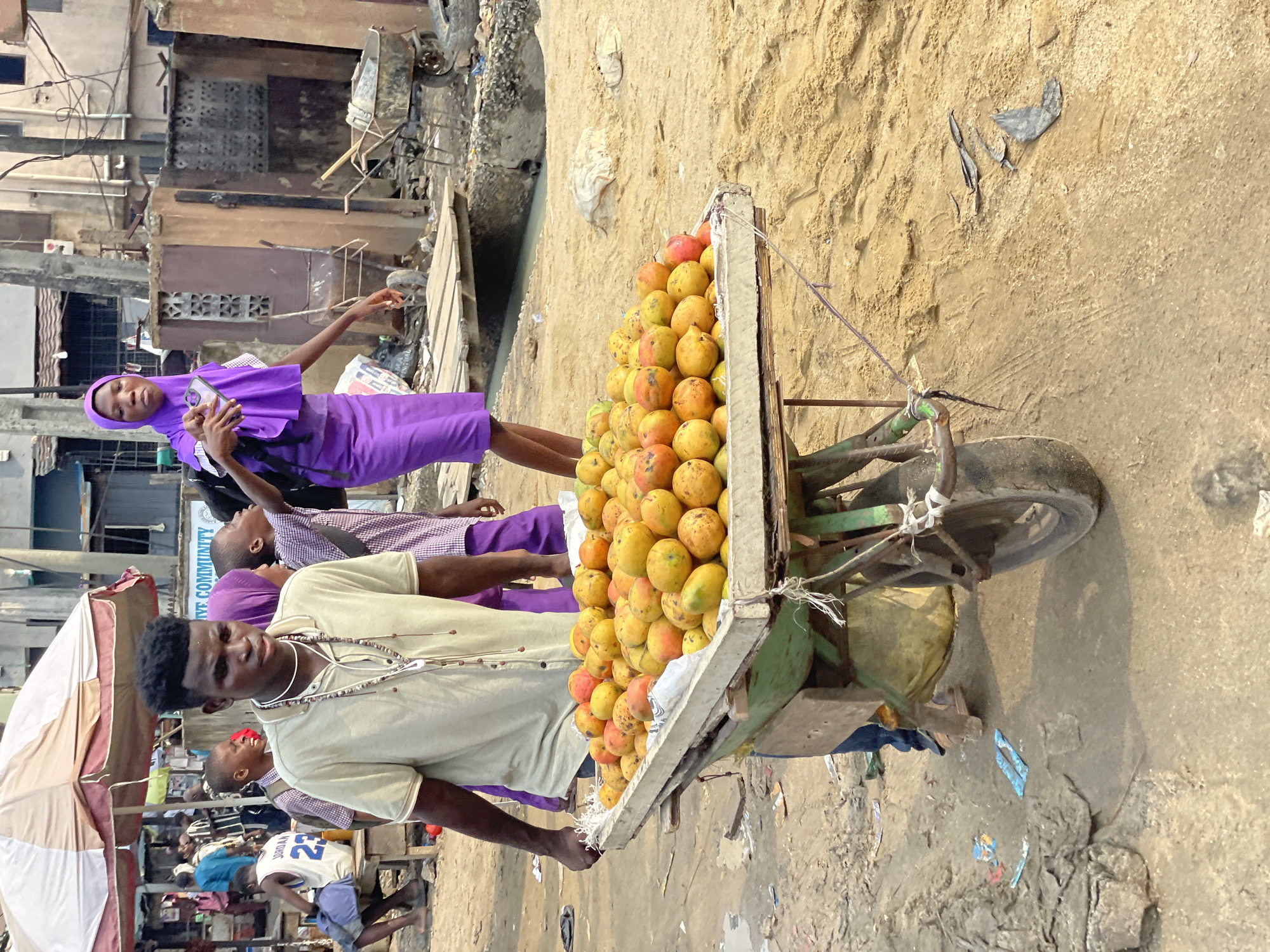 The image shows a man standing next to a wheelbarrow overflowing with mangoes on a street in what appears to be Lagos, Nigeria. The man is African, wearing a light-colored shirt and beaded necklaces. Behind him, another man and a woman in purple clothing are visible, with the woman using a phone. In the background, there is a crowded street scene with buildings and people. The environment suggests a busy, urban marketplace.