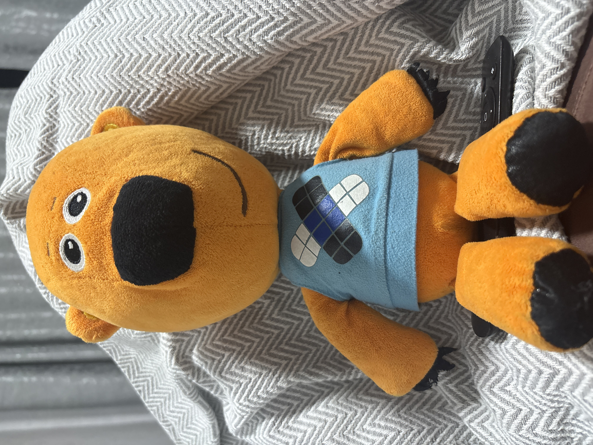 An orange plush bear toy with a blue vest sits on a gray chevron patterned blanket. The bear has a black nose and black paws with visible stitching. A black remote control rests to the right of the bear's feet. The background consists of blurred curtains.