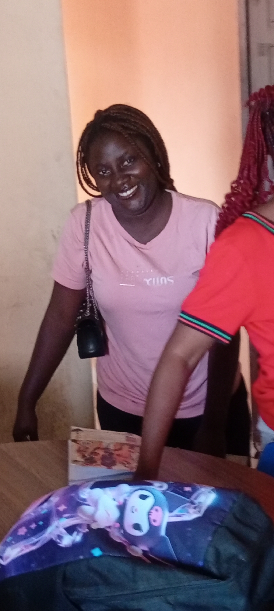 The image shows a woman with braided hair, wearing a pink t-shirt and a black purse, smiling. Another person wearing a red t-shirt is standing to her right. In the foreground, a backpack with a cartoon character is visible on a wooden surface. A box sits on the surface near the woman. The location appears to be indoors. The specific location in Fagge C, Nigeria is not discernible from the image.