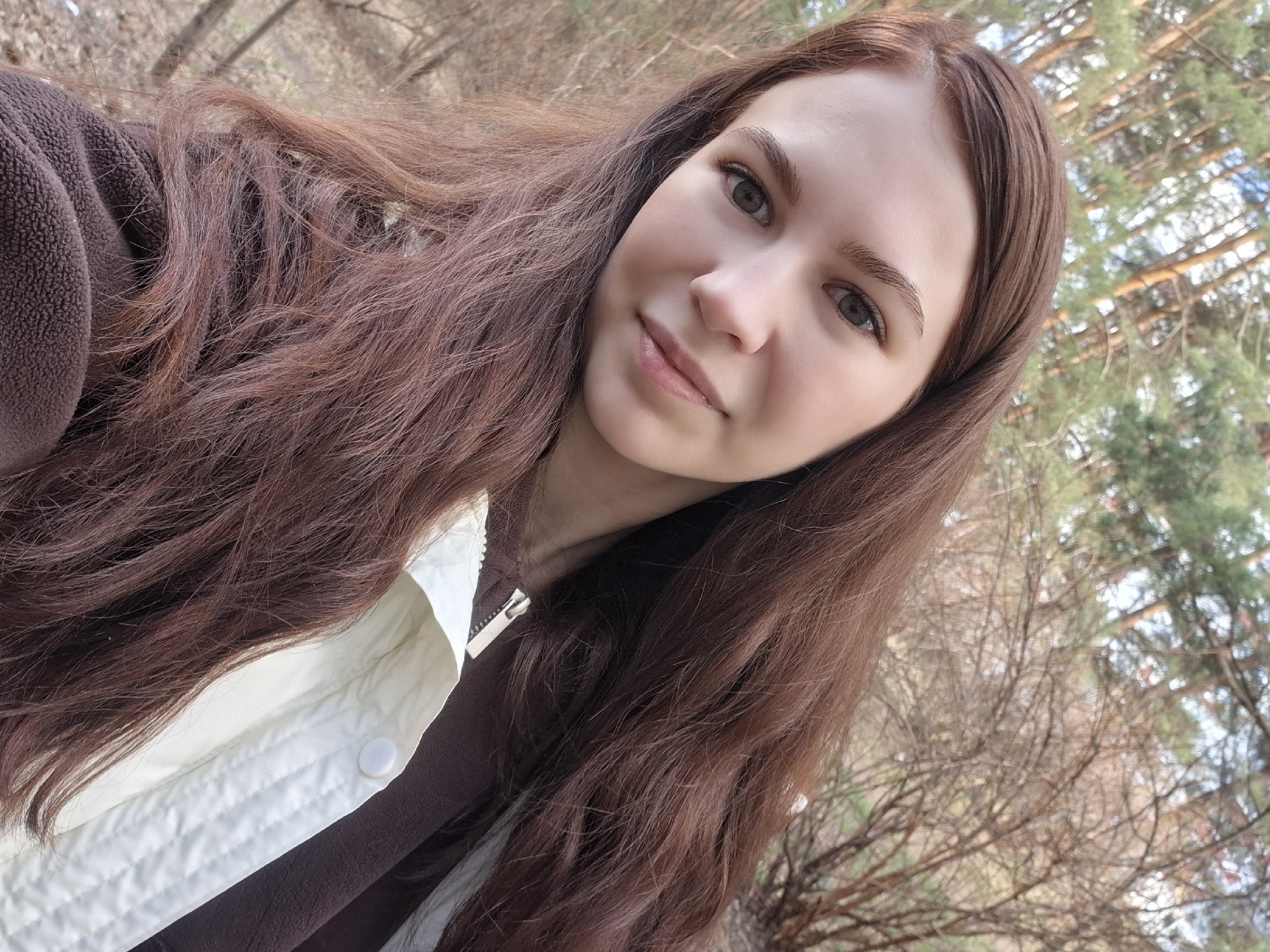 A young woman with long, wavy brown hair is posing for a selfie outdoors. She is wearing a brown fleece jacket under a light-colored jacket with a visible zipper and button. The background shows a natural environment with trees and branches, suggesting she is in a park or wooded area. The photo appears to be taken during the daytime. The location is Chaikovsky, Russia.