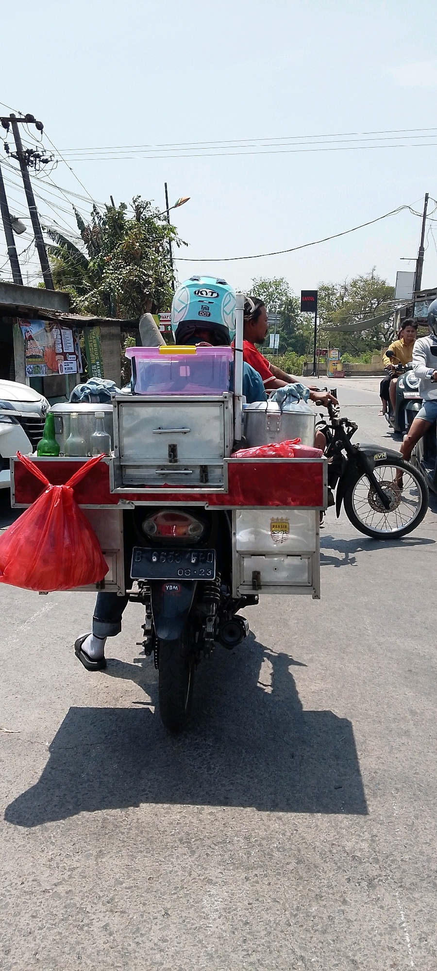 An outdoor, eye-level, medium shot captures a person on a motorcycle in Srimahi, Indonesia, with a cargo carrier attached. The motorcycle's license plate reads "B 6653 FFQ 08-23". The person, wearing a blue and white helmet with "KYT" and "DOT" markings, is seated and facing forward, dressed in a red shirt and blue jeans. A large red plastic bag is prominently displayed on the left side of the cargo carrier, which is laden with various containers, including a translucent pink plastic bin and silver metal boxes. The environment appears to be a street with a paved surface, and the background shows trees and utility poles with wires. The sun is casting a distinct shadow of the motorcycle and its cargo onto the road. Two other motorcycles with riders are visible in the background, suggesting traffic.