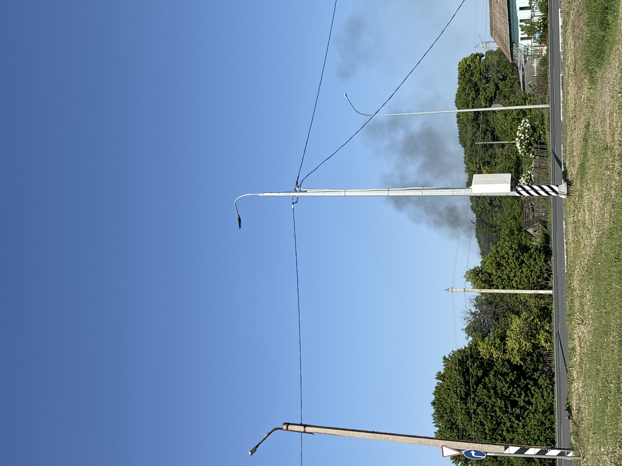 The image depicts a scene in Mar'evka, Russia, showing a roadside view under a clear blue sky. Power lines and streetlights stretch across the frame. In the background, there's vegetation, including trees and some residential buildings. A black and white striped post is visible on the right side of the road. Smoke is also present in the background.