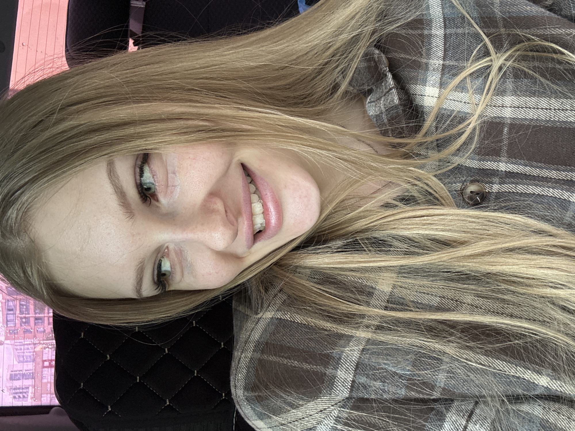A young woman with long, blonde hair is smiling slightly at the camera. She is wearing a brown and grey plaid shirt. She appears to be sitting in a car with a black leather quilted seat. The background shows what might be buildings and a pink/red light reflection. The photo was likely taken indoors, possibly within Perm, Russia given the user's location.