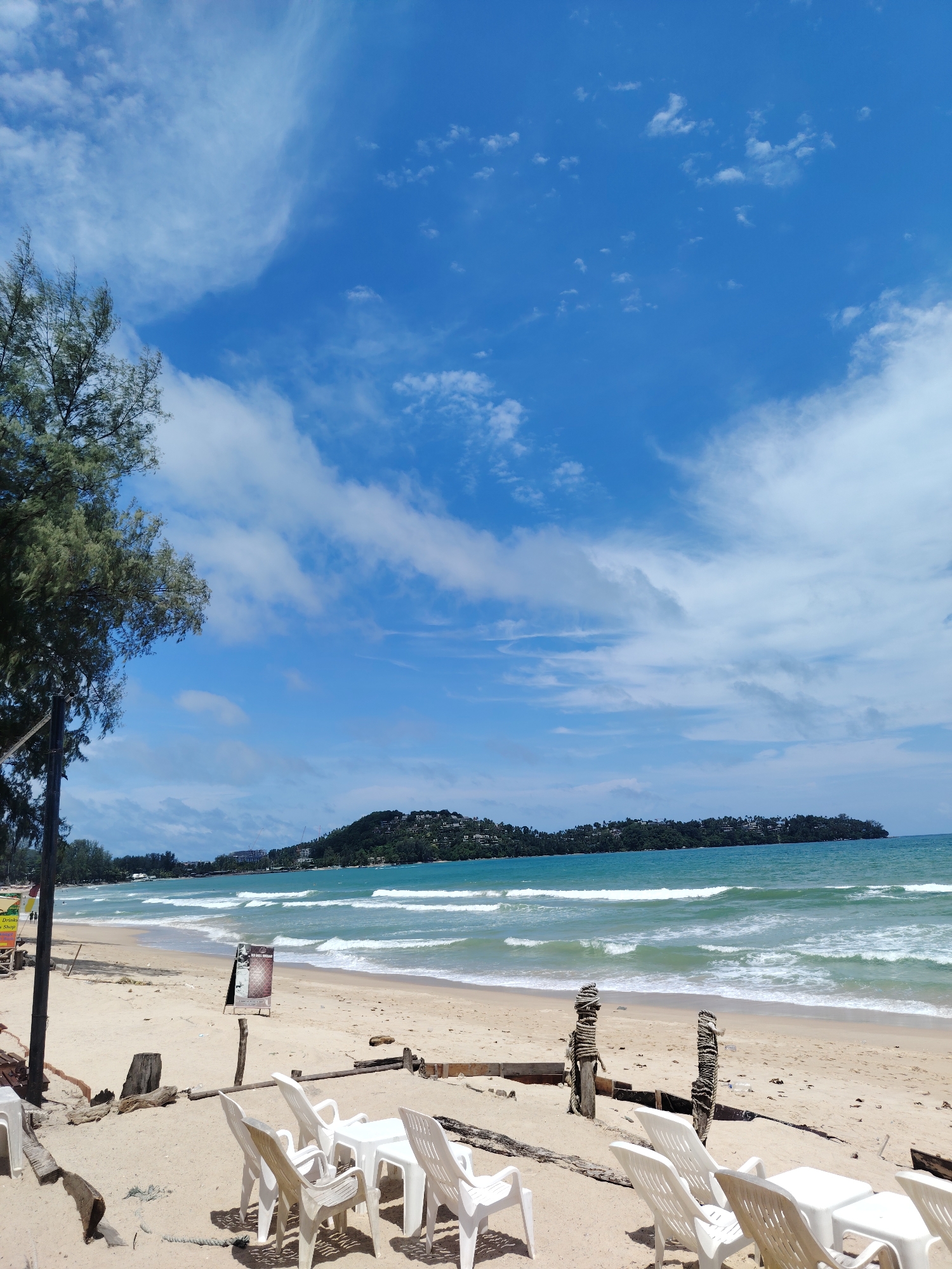 The image depicts a beach scene in Choeng Thale, Thailand. In the foreground, several white plastic chairs are arranged around small white tables on a sandy beach. Some of these chairs and tables are in shadow. To the left, a wooden pole with a sign is visible, and further left, a tree with green foliage extends into the frame. The beach stretches towards the ocean, where waves are breaking. In the mid-ground, there is a dark signpost. In the background, a densely vegetated hill or island rises from the turquoise water, with some buildings visible on its slopes. The sky is a bright blue with scattered white clouds. The overall scene is a bright, sunny day at a tropical beach.