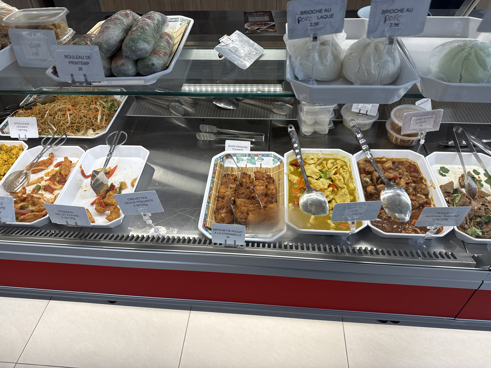 The image displays a deli counter in a food establishment, possibly a grocery store or a restaurant, located in Domont, France. The counter showcases a variety of prepared Asian cuisine offerings. Visible items include spring rolls, noodles with vegetables, seafood dishes (such as shrimp and fish with sauces), chicken skewers, chicken curry, and "Poulet Imperial Piquant" (spicy imperial chicken). There are also bread items labeled "Brioche au Porc" (Pork Brioche) and "Brioche au Porc Laqué" (Lacquer Pork Brioche). Pricing is displayed per item or per 100 grams. Serving utensils are strategically placed within each container, indicating the food is available for immediate purchase.