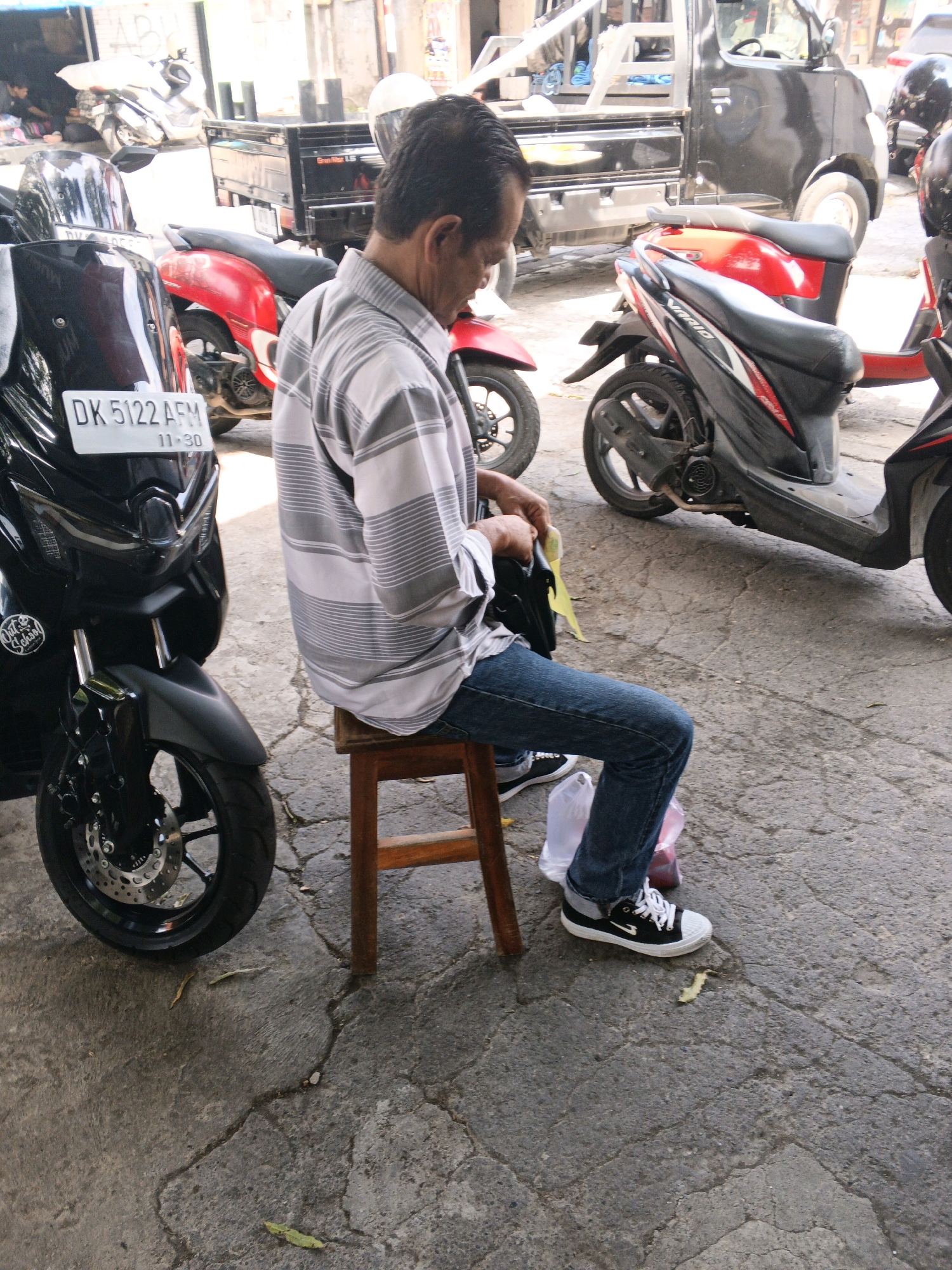 The image shows a man sitting on a low wooden stool on a cracked, grey concrete ground surface, in an outdoor urban environment. He is wearing a grey and white horizontally striped long-sleeved shirt and blue jeans. The man is holding a black wallet or bag in his left hand and a yellow piece of paper in his right hand. He is wearing black and white sneakers.
Behind the man and to his left, a black motorcycle with a white license plate (DK 5122 AFM) is parked. The motorcycle has a windshield and is partially obscured by the man. To the right of the man and behind him, another motorcycle with a red seat and body is parked. Further behind, there is a black truck with a wooden flatbed. In the background, there are hints of buildings and structures, suggesting a street or marketplace setting. The lighting suggests it is daytime. The overall scene is one of everyday life in a busy urban area. Given the license plate format and style of vehicles, this scene is consistent with being in Denpasar, Indonesia.