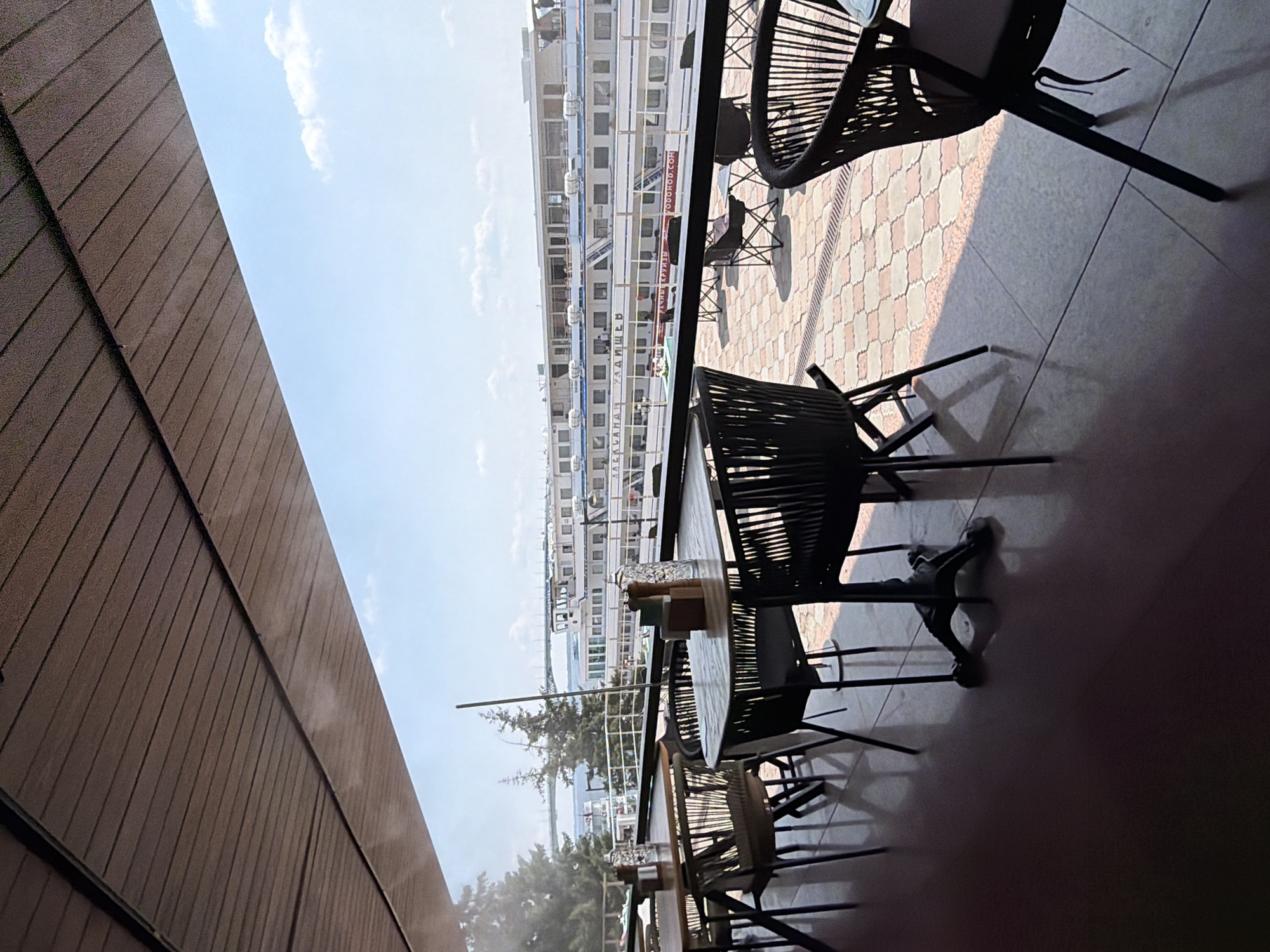 The image depicts an outdoor seating area of a restaurant or cafe, likely located in Saratov, Russia. The view extends to a waterfront where a large, multi-deck passenger ship is docked. The ship is white with multiple rows of windows and appears to be named "АЛЕКСАНДРА РАДИЩЕВ". In the background, a bridge is visible spanning the water, suggesting a river or large body of water.
In the foreground, several tables and chairs are arranged on a patterned tiled patio. The tables are wooden with metal legs, and some are set with salt and pepper shakers and water bottles. The chairs are modern, black wicker-like material, some with cushions. The seating area is partially covered by a wooden-slatted awning. The overall atmosphere is bright and sunny, with a clear blue sky and some scattered clouds.
There are no people visible in the foreground seating area, but there appear to be a few individuals on the deck of the ship. The scene suggests a pleasant day for outdoor dining with a scenic view of the waterfront and river traffic.
