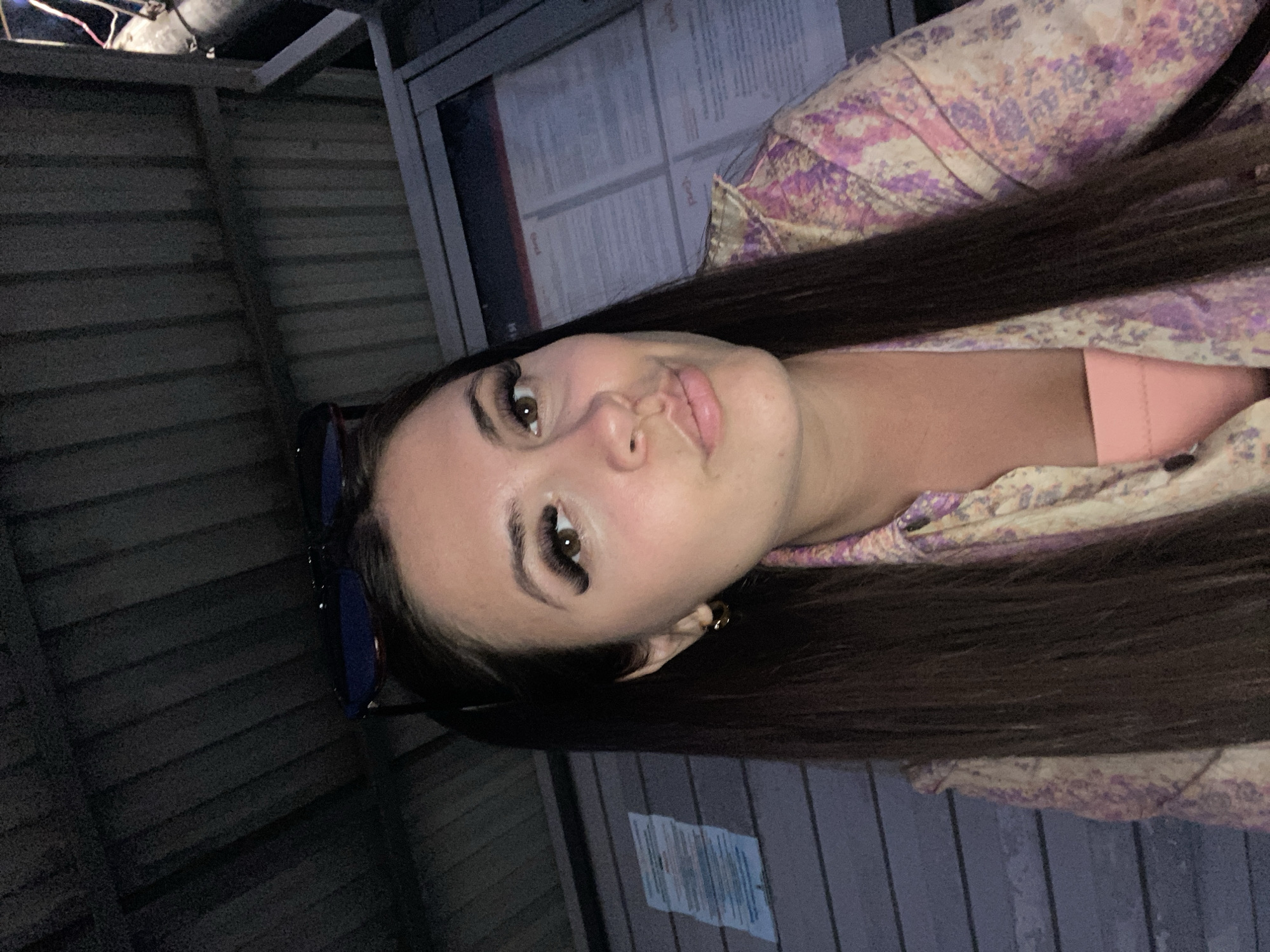 In Samur, Russia, a young woman with long, dark hair and dramatic false eyelashes is pictured in a close-up selfie. She is wearing a colorful patterned shirt over a pink garment, and a pair of sunglasses rests on her head. The background appears to be an outdoor covered area, possibly a bus stop or waiting shelter, with corrugated metal roofing and siding. A notice board or informational display is visible behind her. The lighting suggests it is nighttime or a dimly lit environment, with some light sources creating highlights on her face and the environment.