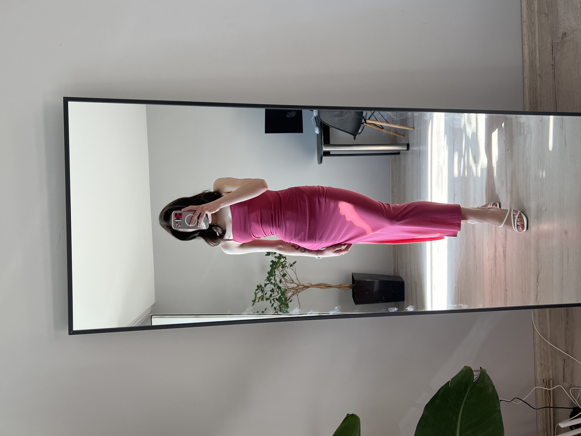 A person stands in front of a full-length mirror, taking a picture of their reflection. They are wearing a form-fitting, strappy, hot pink maxi dress with a slit on the side, and white strappy heeled sandals. The person has dark hair and is holding a smartphone in their right hand, using it to capture the image. In the reflection, a potted plant with green leaves sits to the left of the person. Behind the plant, a dark object, possibly a computer monitor, is visible on a small table with a black chair. The room has light gray walls and light-colored wooden flooring. The scene is lit by natural light, casting shadows on the floor. The image appears to be taken indoors, likely in a residential setting.