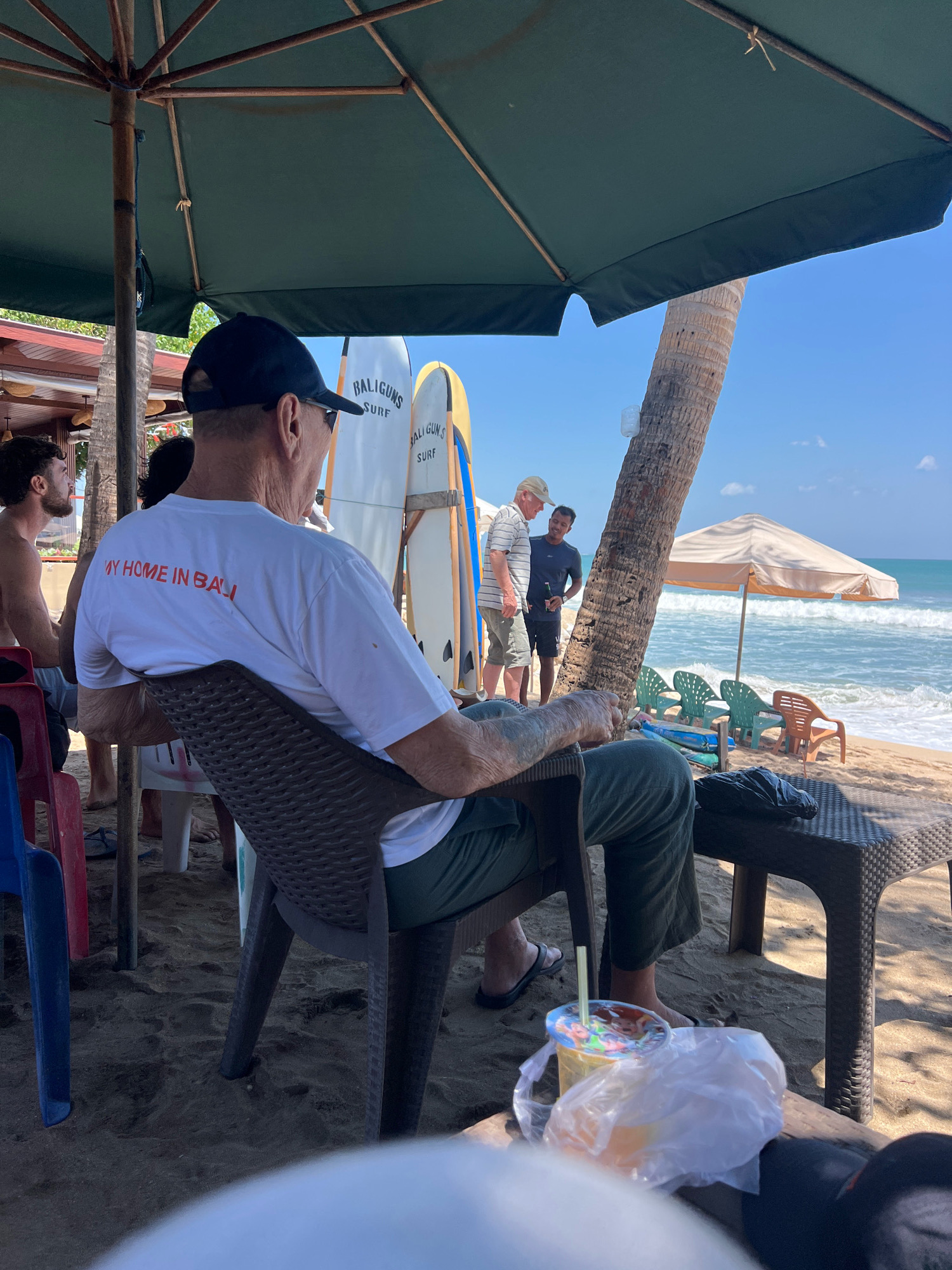 An outdoor, eye-level, medium shot captures a beach scene in Kuta, Indonesia, under a large green umbrella. In the foreground, a man wearing a white t-shirt with the text "MY HOME IN BALI" and green pants sits on a dark brown plastic chair, facing away from the camera. A plastic cup with a yellow beverage and a straw rests on a small table to his right, partially covered by a white plastic bag. Behind him, two surfboards labeled "BALIGUNS SURF" stand upright. In the mid-ground, two men are conversing near a palm tree. One man, wearing a striped shirt and a light-colored hat, appears to be speaking to another man in a dark t-shirt. Further back, several empty green plastic chairs and one orange plastic chair are arranged on the sand, facing the ocean. The background features the blue ocean with gentle waves and a clear sky with scattered clouds. The overall atmosphere suggests a relaxed beach setting.
