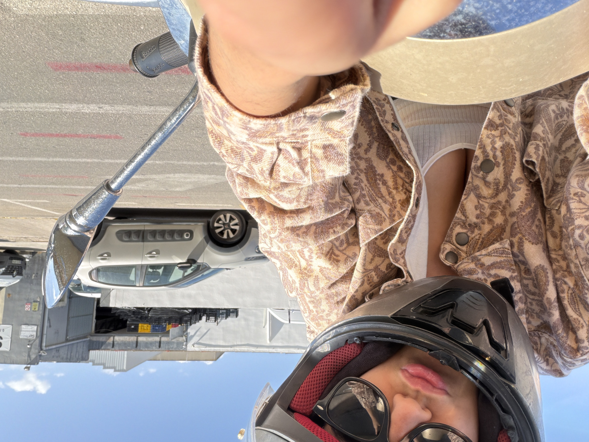 The image captures a selfie-style, low-angle perspective of a person on what appears to be a motorcycle or scooter, set against an outdoor urban or industrial backdrop under bright daylight.
The main subject is a person partially visible in the foreground, wearing a black motorcycle helmet with red interior padding and dark sunglasses. Their lips, tinted pink, are visible below the helmet visor. They are wearing a light brown or beige patterned jacket with buttons, possibly a denim-like fabric, over a ribbed, light-colored (likely white or cream) tank top or undershirt. A bare arm or forearm is also visible, positioned near the vehicle's controls.
Parts of the motorcycle/scooter are prominent: a chrome handlebar or mirror stem extends into the frame, and a black rubber grip with a textured pattern is visible, faintly displaying the brand name "Vespa".
In the mid-ground and background, the scene reveals an asphalt surface with painted red and white lines, suggesting a road or parking area. A grey modern car, resembling a Citroën C3 Aircross with black alloy wheels, is parked. Beyond the car, various industrial or commercial buildings with grey walls and some corrugated metal siding are present under a clear blue sky with scattered white clouds. A stack of black plastic crates is visible near one of the buildings. Small signs are present on the buildings, but their text is unreadable. The overall setting suggests a sunny day in an urban or industrial environment.