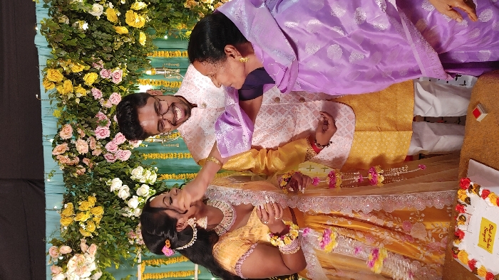 This image captures a heartfelt moment from a Sangeet ceremony, a pre-wedding celebration, taking place in Dhaka, Bangladesh. The scene features three individuals amidst a vibrant, festively decorated setting.
In the foreground, a young woman, likely the bride, is dressed in an ornate yellow and gold embellished outfit, adorned with traditional floral jewelry, including gajras on her wrists. An older woman, wearing a light purple saree or dupatta, is affectionately feeding a bite of food, possibly cake or a sweet, to the young woman. The young woman looks down with a subtle smile, accepting the gesture. To the left, a young man, presumed to be the groom, stands smiling broadly at the camera. He wears glasses and a light-colored, intricately embroidered jacket.
The background is lush with extensive floral decorations, featuring a vibrant arrangement of yellow, pink, and white roses intermingled with green foliage, creating a beautiful backdrop. Additional yellow garlands are draped across a teal blue fabric, adding to the festive atmosphere. The setting appears to be an indoor event, brightly lit, suggesting a joyous celebration.
On the right side of the image, a white cake decorated with yellow and red floral icing is visible. A label on the cake clearly reads, "Badh O Bodhir Sangeet," confirming the event's nature as a Sangeet for the bride and groom. A small red and white box is also seen near the cake. The overall mood is one of warmth, happiness, and familial affection, characteristic of such celebratory events.