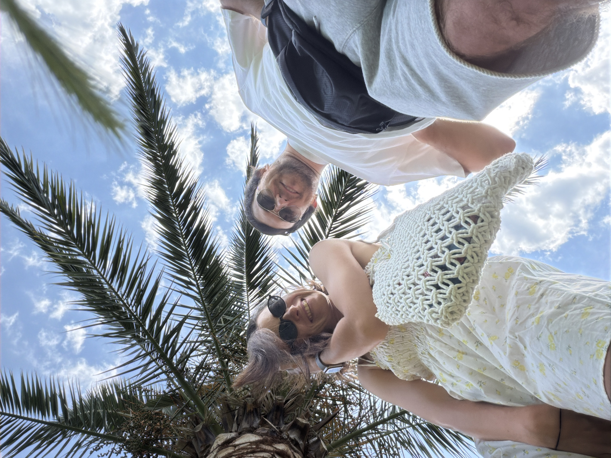 Two individuals, a woman and a man, are captured from a low-angle perspective, looking up towards a bright, partly cloudy sky, with palm fronds overhead. The woman, on the left, wears sunglasses and a light-colored floral dress. She is smiling and holding a woven white bag. Her hair is windswept. The man, on the right, also wears sunglasses and a white t-shirt, with a black fanny pack across his chest and light-colored shorts. They appear to be outdoors in a warm climate, potentially near a beach or park. The image was taken in Budva, Montenegro.