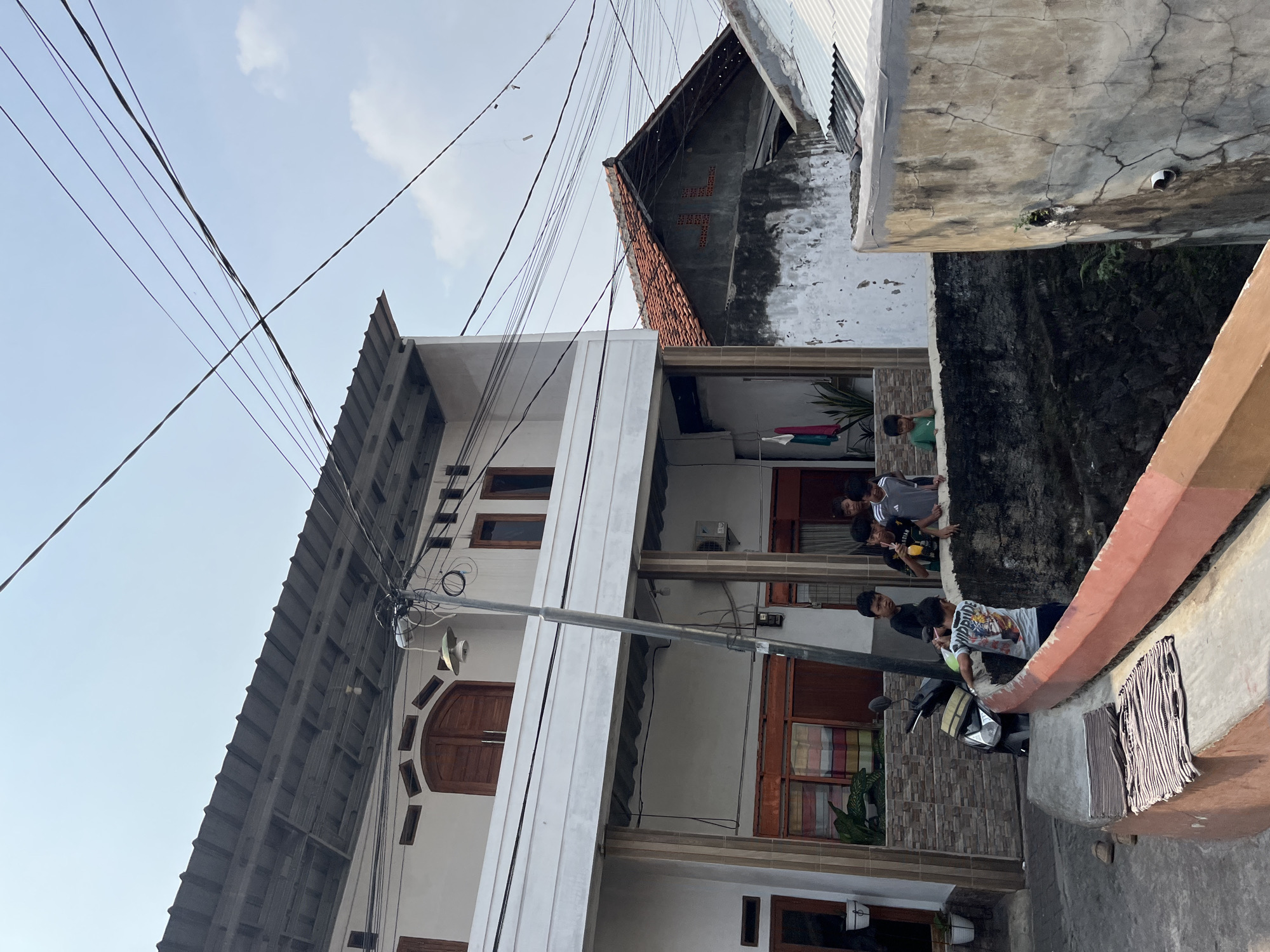 The image depicts a street scene in Tangerang, Indonesia, likely a residential area, characterized by a mix of buildings and overhead electrical wires. In the foreground, a concrete barrier with an orange railing separates the viewer from a lower level, where several boys are gathered. A motorcycle is parked to the left of the frame.
The central focus is a two-story building with a white facade and dark gray metal roofing. The upper floor has a balcony with wooden framed windows. The lower floor features pillars, stone-like cladding, and windows with colorful curtains. A street lamp on a pole stands prominently near the building.
To the right of the main building, another structure with a red tiled roof is visible, showing signs of age and some disrepair. More electrical wires crisscross the sky, indicating an urban or semi-urban setting.
The group of boys, appearing to be between 8 and 15 years old, are interacting near the barrier. Some are looking towards the camera, while others seem engaged in conversation or observing something in the vicinity. One boy in the foreground, wearing a patterned t-shirt, is leaning over the barrier. Another boy, in a grey shirt, holds a drink. A younger boy in a green shirt is visible on the right side of the barrier. The presence of laundry hanging from a line on the building suggests an inhabited area. The overall impression is a snapshot of everyday life in a bustling neighborhood.