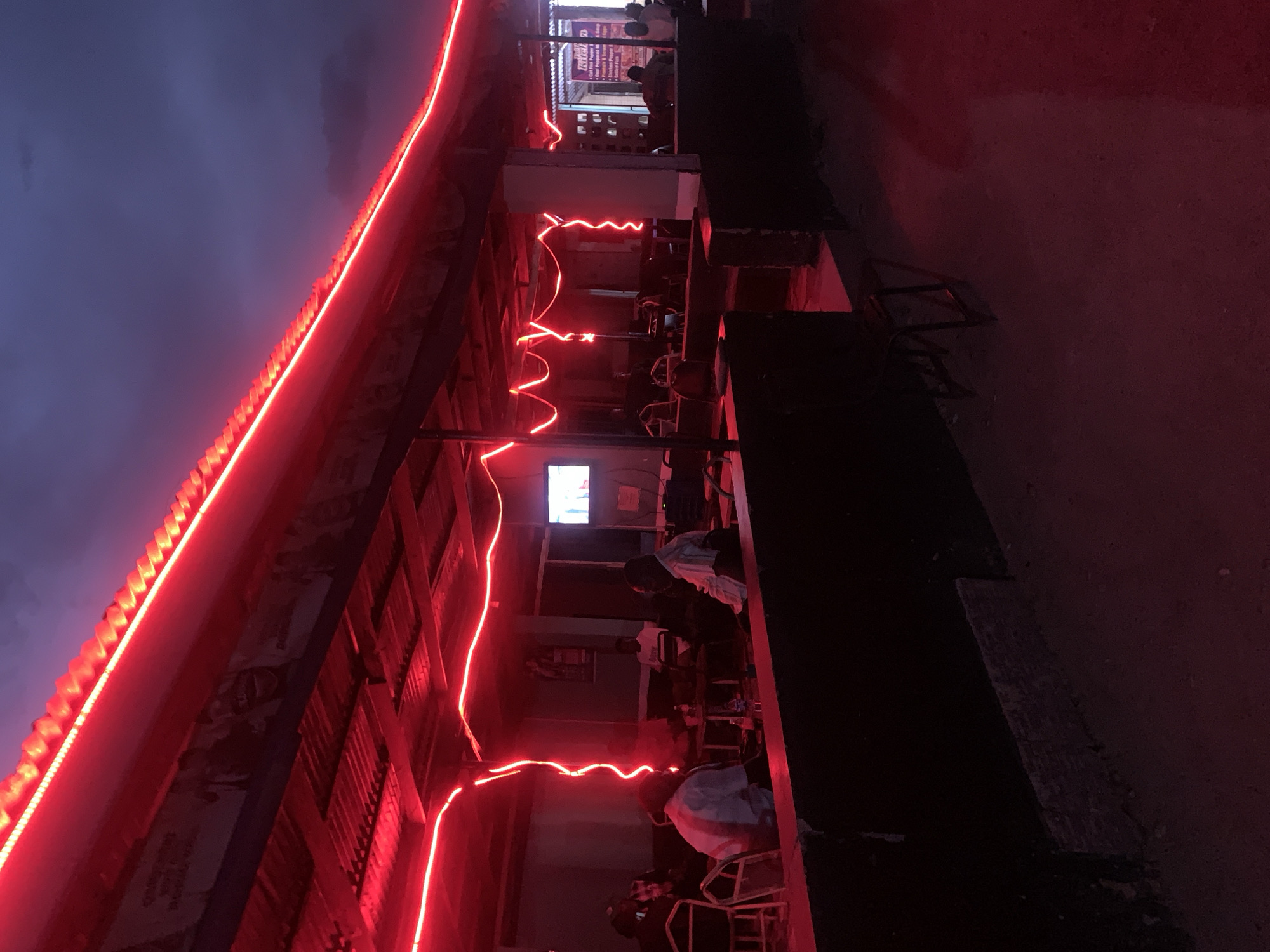 The image shows an outdoor area in the evening, possibly at or near a building in the New Stadium area of Nigeria. The scene is illuminated by red neon lights outlining the building's structure. Several people are seated, seemingly patrons of the establishment, engaged in conversations or watching television. The environment is dark, with limited ambient lighting, creating a shadowed effect. A dirt road is adjacent to the building.