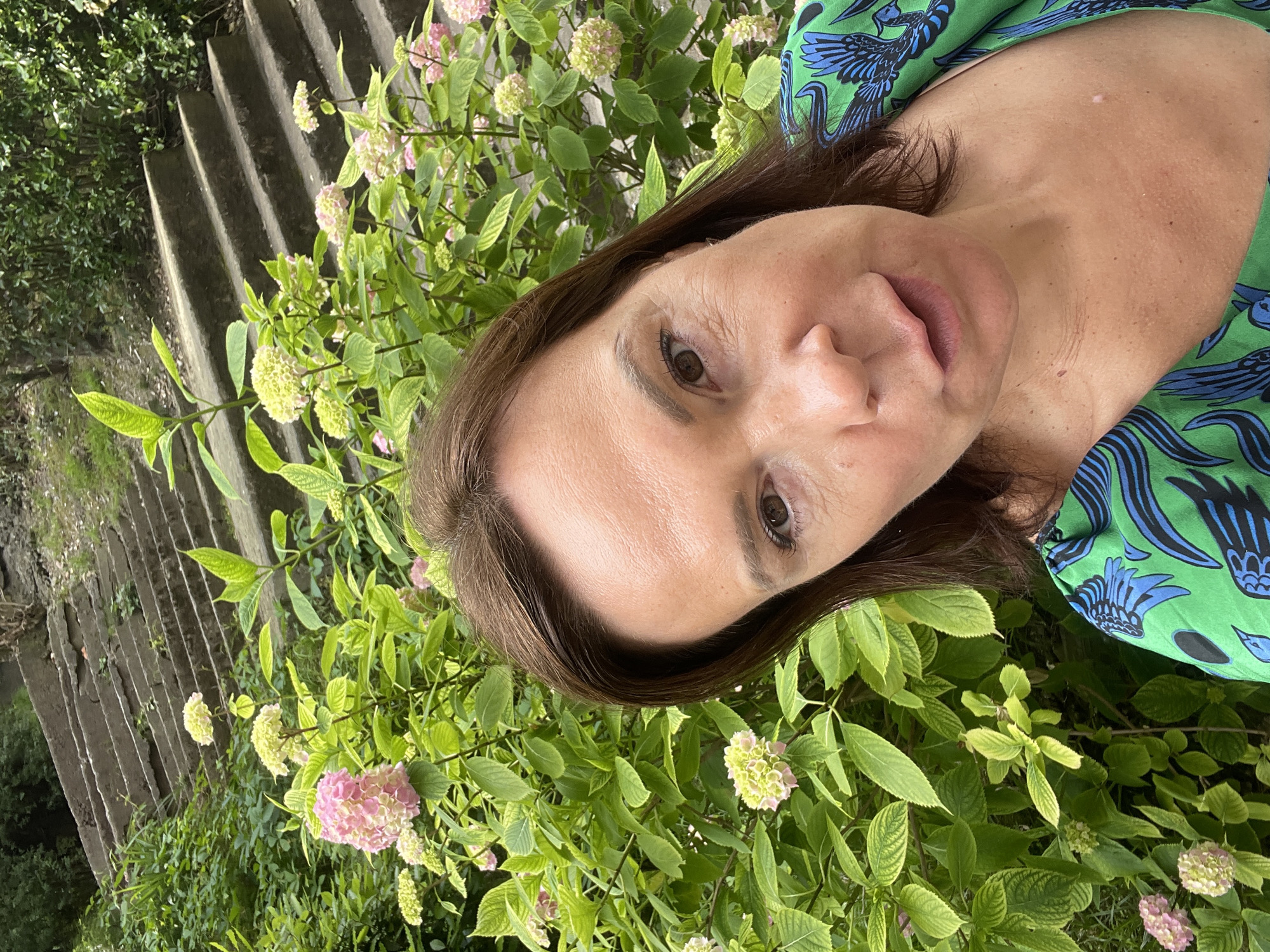 A woman is taking a selfie in front of a dense green bush with pink and white flowers, and a concrete staircase in the background. She has brown hair and is wearing a green dress with a blue bird pattern. The environment appears to be an outdoor setting with lush vegetation and a weathered staircase. The location is identified as Sochi, Russia.