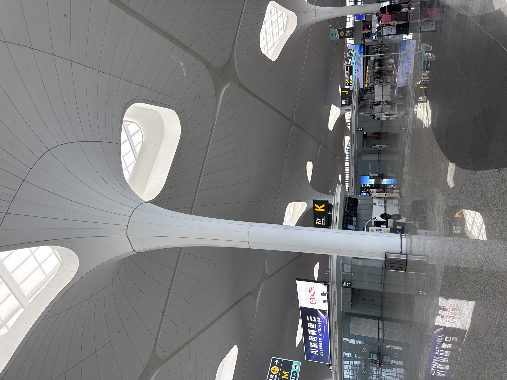 This image depicts the interior of a modern airport terminal, likely Xiaoshan International Airport in Xiaoshan District, China, given the context. The architecture is characterized by a large, undulating ceiling with a grid-like pattern and strategically placed skylights that allow natural light to illuminate the space. Several large, white, sculptural columns support the ceiling, resembling stylized trees.
The floor is highly polished, reflecting the overhead lighting and architectural features. Various directional signs are visible, indicating check-in areas, gates, and possibly international and domestic departures. One prominent sign displays "AI just use Alibaba Cloud" with Chinese characters. Another sign indicates "K" and "International Hong Kong, Macau, Taiwan." A digital display board shows flight information.
Several people are present in the terminal, walking or standing. Some are near check-in counters or information kiosks. A white utility vehicle is parked in the background. The environment appears clean, spacious, and functional, consistent with a busy transportation hub. The overall impression is one of contemporary design and efficiency.