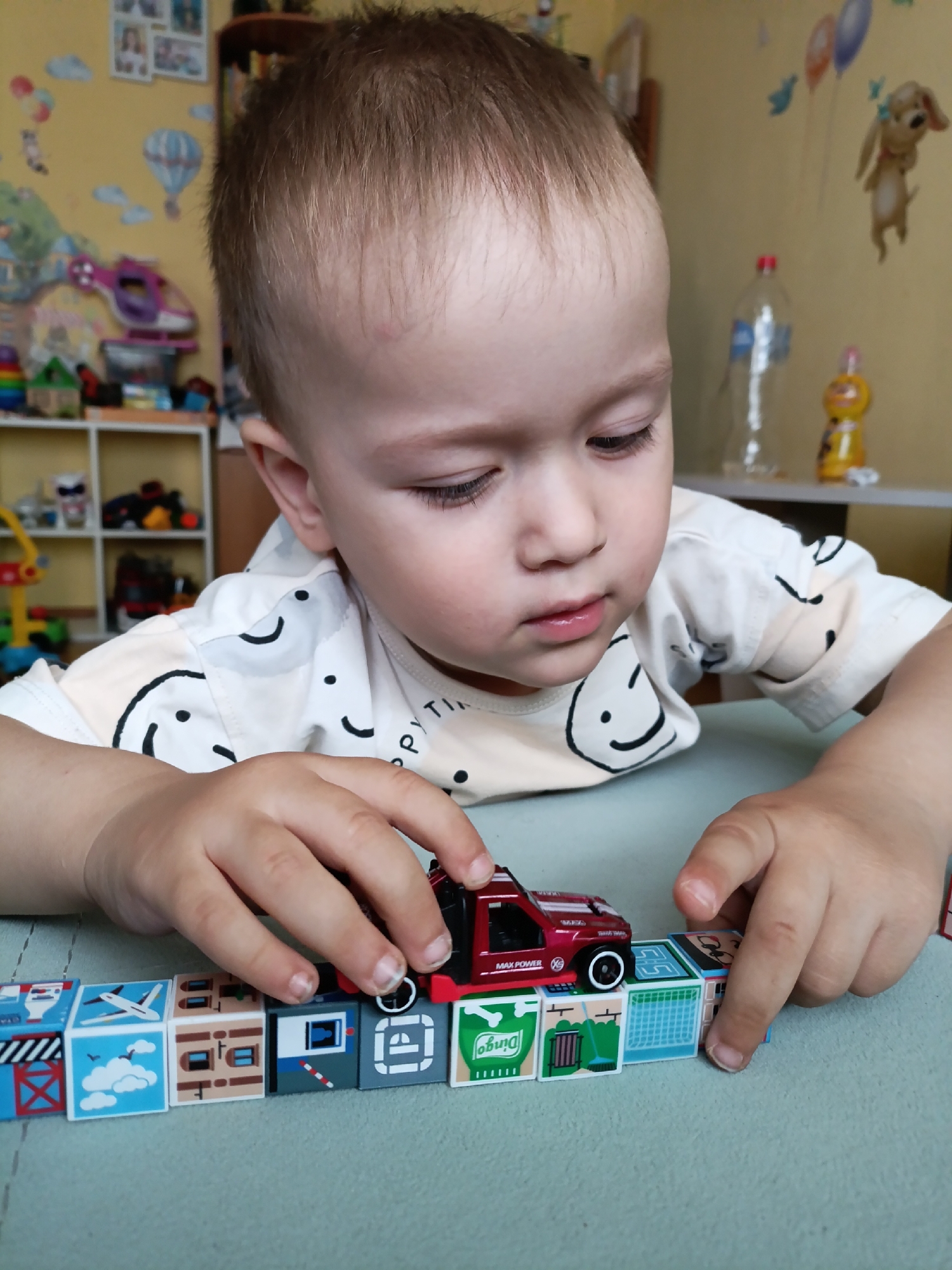A young child, with light brown hair and fair skin, is seated at a table in what appears to be a domestic setting. The child is focused on a line of toy blocks laid out on the table. A small red toy truck is positioned on top of two of the blocks. The child's hands are actively engaged with the toys. The background suggests a child's room, with colorful decals of balloons, a hot air balloon, and a dog on a yellow wall. Shelving units filled with various toys and items are visible in the background. The lighting indicates an indoor environment, likely during the day. The image does not provide any specific information to determine the location within Chaikovsky, Russia.