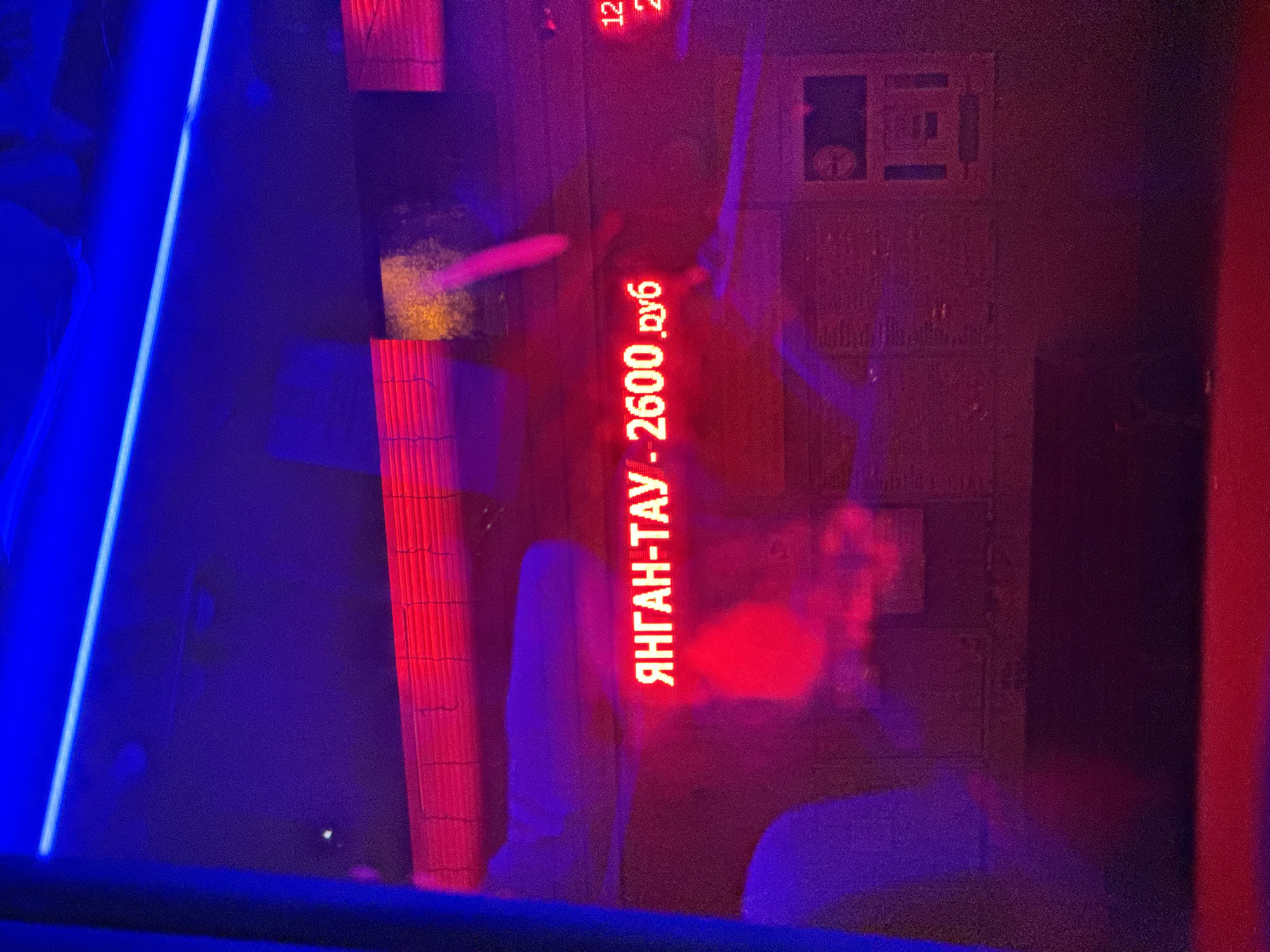 A neon sign in Russian reads "ЯНГАН-ТАУ - 2600 руб", meaning "Yangantau - 2600 rubles". The sign is red and is illuminated by neon lights. The sign is in a dark environment, possibly a bar or nightclub. There are also blue lights in the background. The location is identified as Podgornyi Driush, Russia.