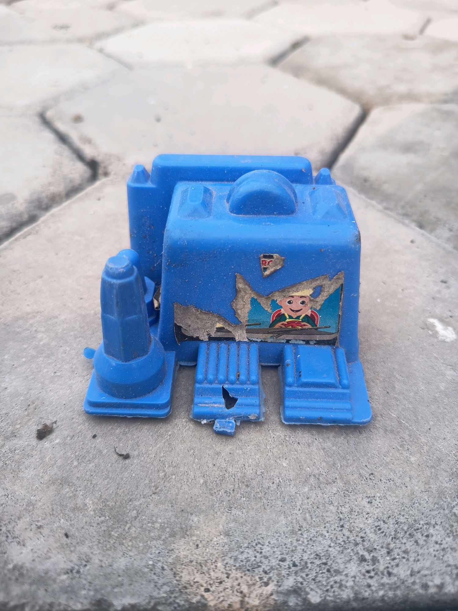 A blue plastic toy truck sits on a gray paved surface. The toy is damaged, with torn stickers revealing a cartoonish driver behind the windshield. The paving stones are arranged in a hexagonal pattern. The scene appears to be outdoors in an urban environment, possibly Sidorejo, Indonesia, given the context. There are no people or animals present.