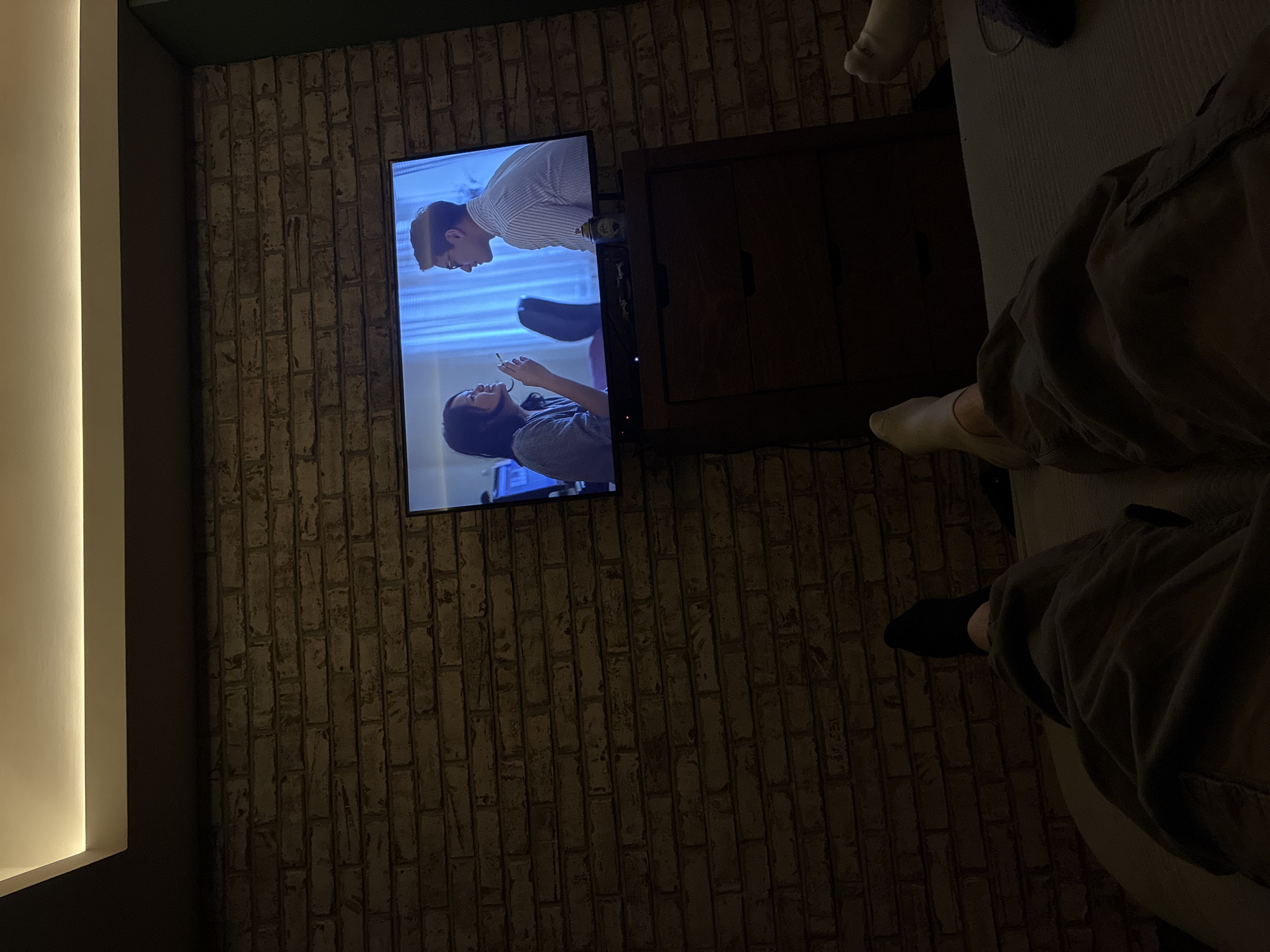 A television is mounted on a brick-patterned wall in a dimly lit room. On the television screen, two figures are visible: a man in a striped shirt looking down at a woman holding a cigarette. A dark wooden cabinet is adjacent to the television. A person is partially visible on the right side of the image, wearing baggy gray pants with a sock on one foot and a black sock on the other. The setting is indoors, and based on the wall's pattern and general ambiance, it may be located in a home. No information about a precise location in Huixquilucan de Degollado, Mexico, is discernible from the image.
