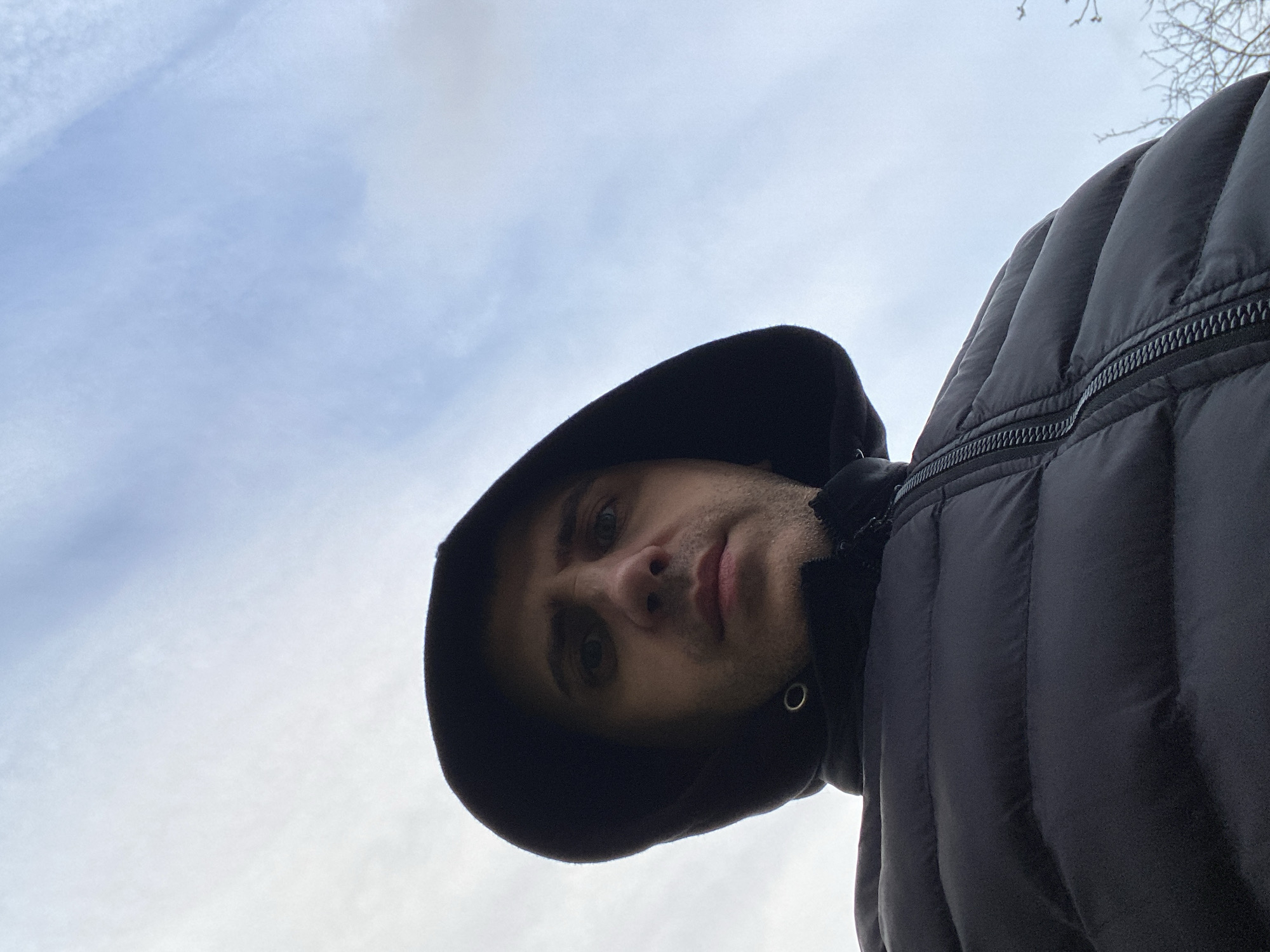 A low-angle, upward-facing photograph captures a person wearing a black hooded puffer jacket. The sky is visible in the background with light, wispy clouds. A few bare tree branches appear in the upper right corner of the frame. The location is Unknown, Unknown.