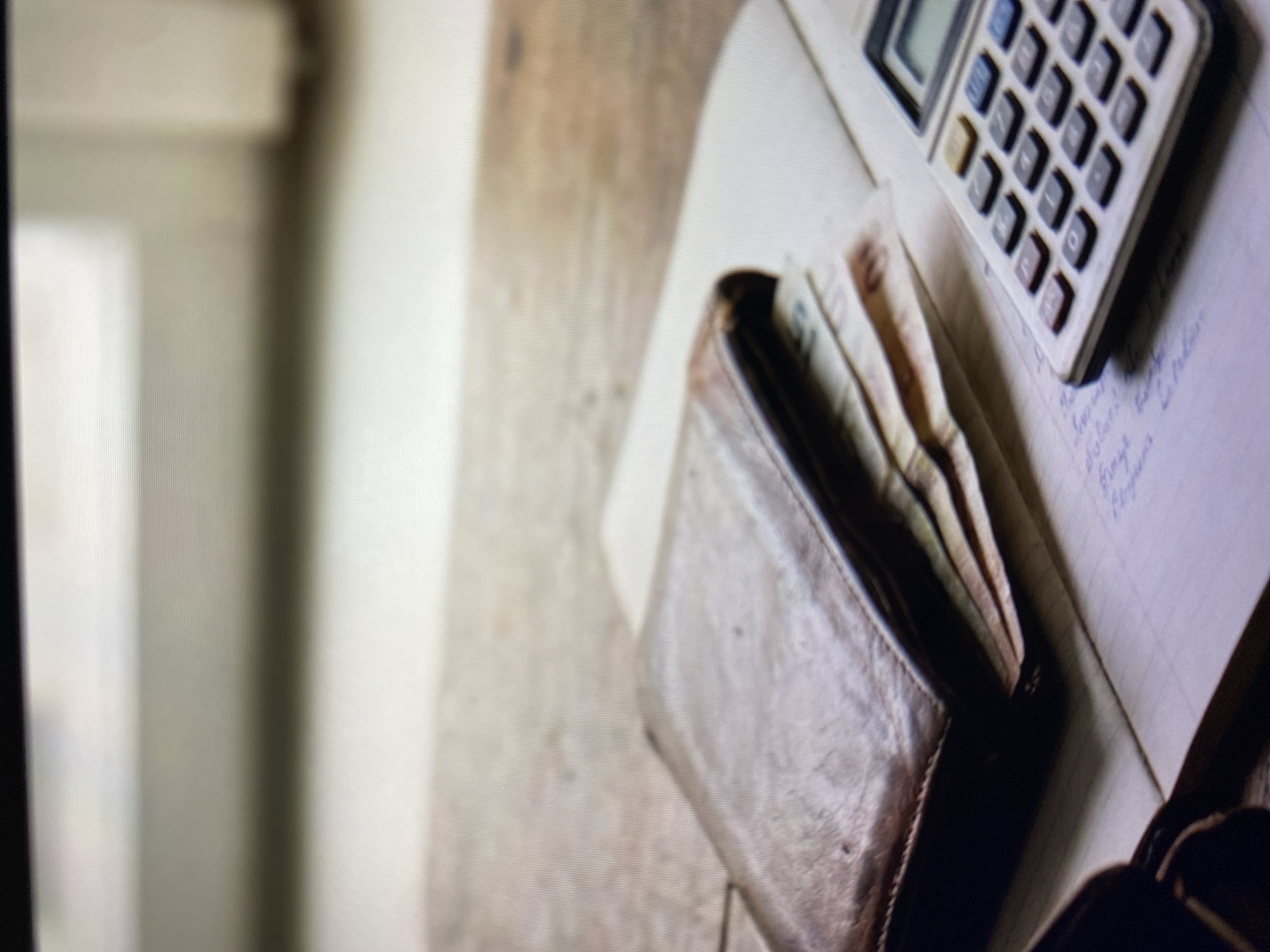 An overhead shot shows a weathered brown leather wallet resting open on a wooden surface, revealing several banknotes tucked inside. To the right of the wallet, a portion of a notebook with faint blue handwritten text is visible, along with a small, grey calculator with multi-colored buttons. The background features a blurry light source, suggesting a window. The scene is indoors, likely a domestic or office setting, and offers no discernible clues related to Kuta Selatan, Indonesia.