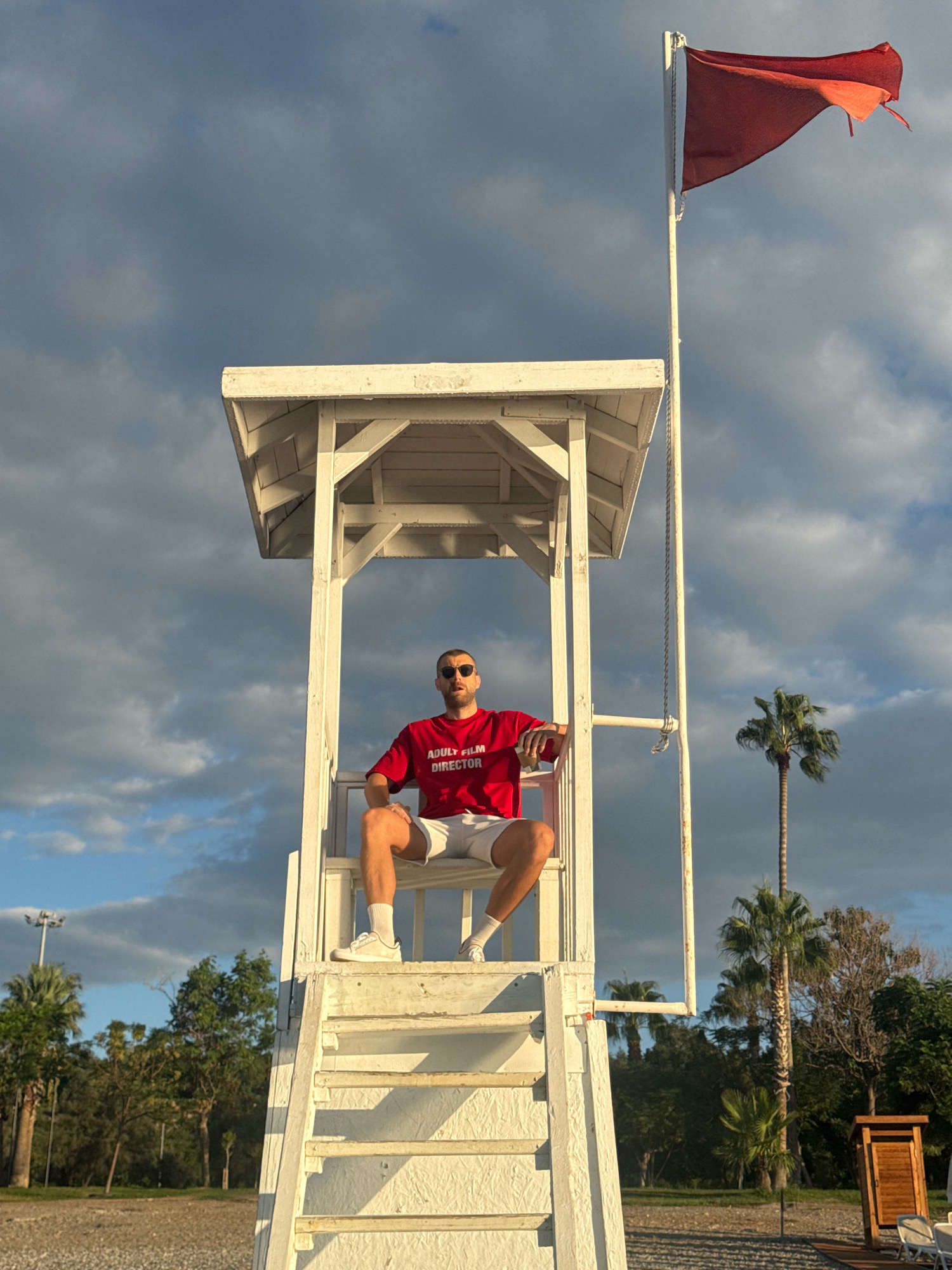 A low-angle, medium shot captures a man seated on a white lifeguard chair on a beach in Antalya, Turkey. The man, wearing sunglasses, a red t-shirt with white text that reads "ADULT FILM DIRECTOR," and white shorts with white socks and sneakers, looks directly at the camera. His legs are crossed, and his left arm rests on his knee. The lifeguard chair is elevated, with a red flag flying from a pole on the right. The background features a cloudy sky, palm trees, and a dimly lit wooden structure. The ground appears to be composed of pebbles.