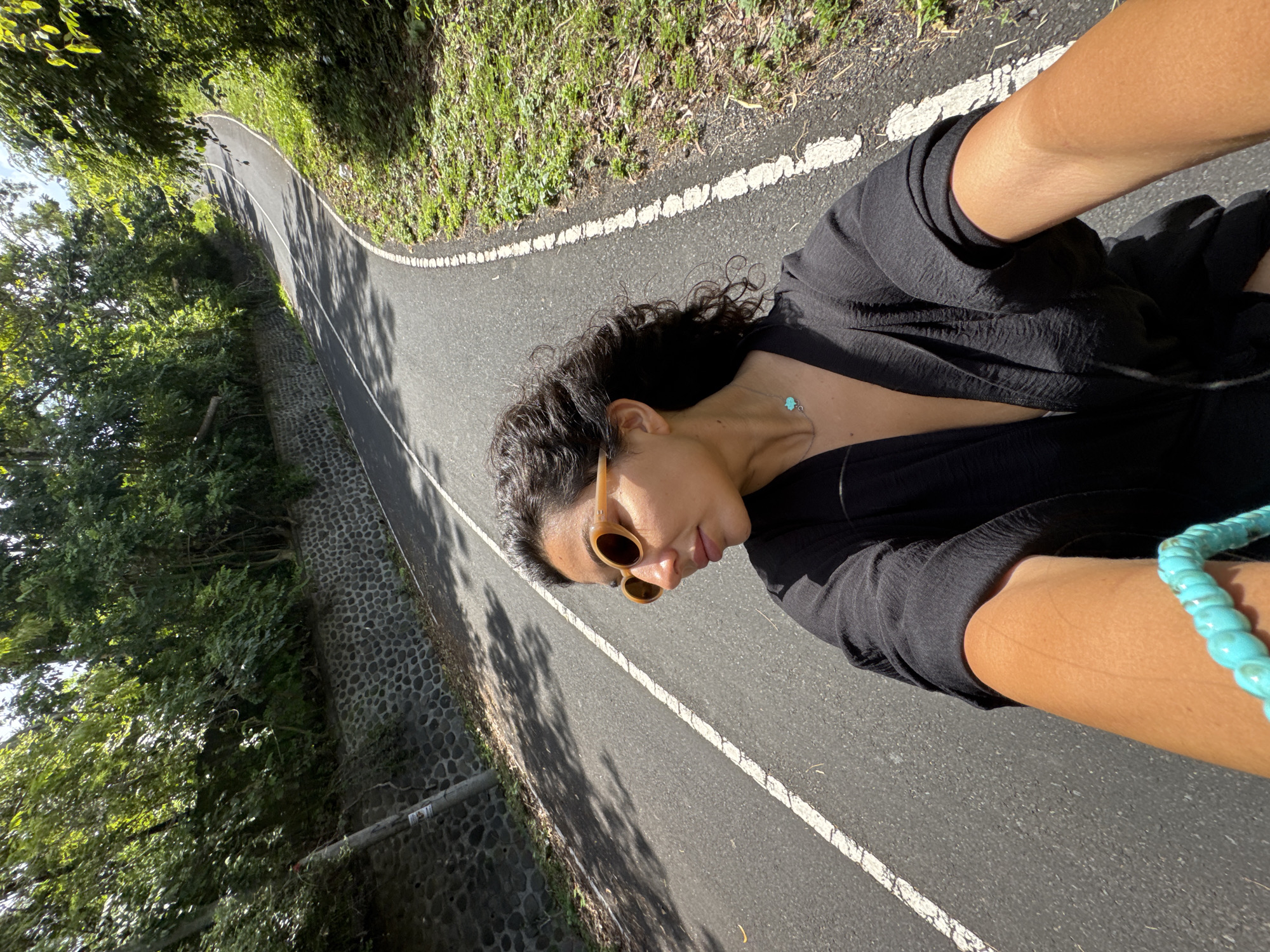 A woman is on a paved asphalt road with a white dividing line, likely a walking path. She's wearing a black top and brown-rimmed sunglasses. A turquoise necklace and a turquoise beaded bracelet are visible. The road is lined with trees and greenery on one side and a stone wall on the other. The scene appears to be outdoors in sunlight. Given the location of Kuta Selatan, Indonesia, it's possible this image was taken in Uluwatu.
