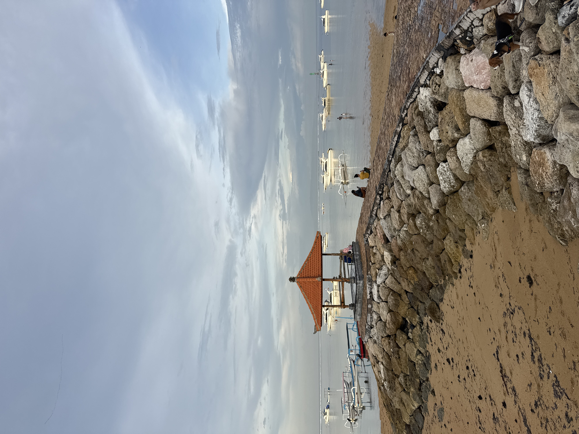 This image depicts a serene coastal scene in Denpasar, Indonesia, under an overcast sky. The overall setting is a beach or bay area featuring calm water, a sandy shore, and a prominent stone seawall.
The upper portion of the frame is dominated by a soft, cloudy sky, suggesting an overcast day with diffused light rather than direct sunlight. Below the sky, a tranquil body of water extends, dotted with numerous small, traditional outrigger boats, likely local Jukungs. These boats, predominantly white with some blue detailing, are anchored in the shallow water, with several people visible standing or wading near them in the distance.
In the left foreground, a small, open-sided gazebo or balé with a distinctive orange-red tiled roof and wooden supports stands on the stone seawall. Two individuals are seated within this shelter, seemingly enjoying the peaceful surroundings. The seawall itself, made from large, rough-hewn stones, curves along the edge of the water, separating it from the sandy shore.
The sandy beach area in the foreground and along the right side of the image shows some darker patches, possibly seaweed or wet sand. Towards the right side of the seawall, a person is seated next to a brown and white dog, which appears to be a beagle, wearing a harness and looking out towards the water. Another person is seated directly on the sand further to the right.
The scene conveys a quiet and relaxed atmosphere. No clear text is legible within the image.