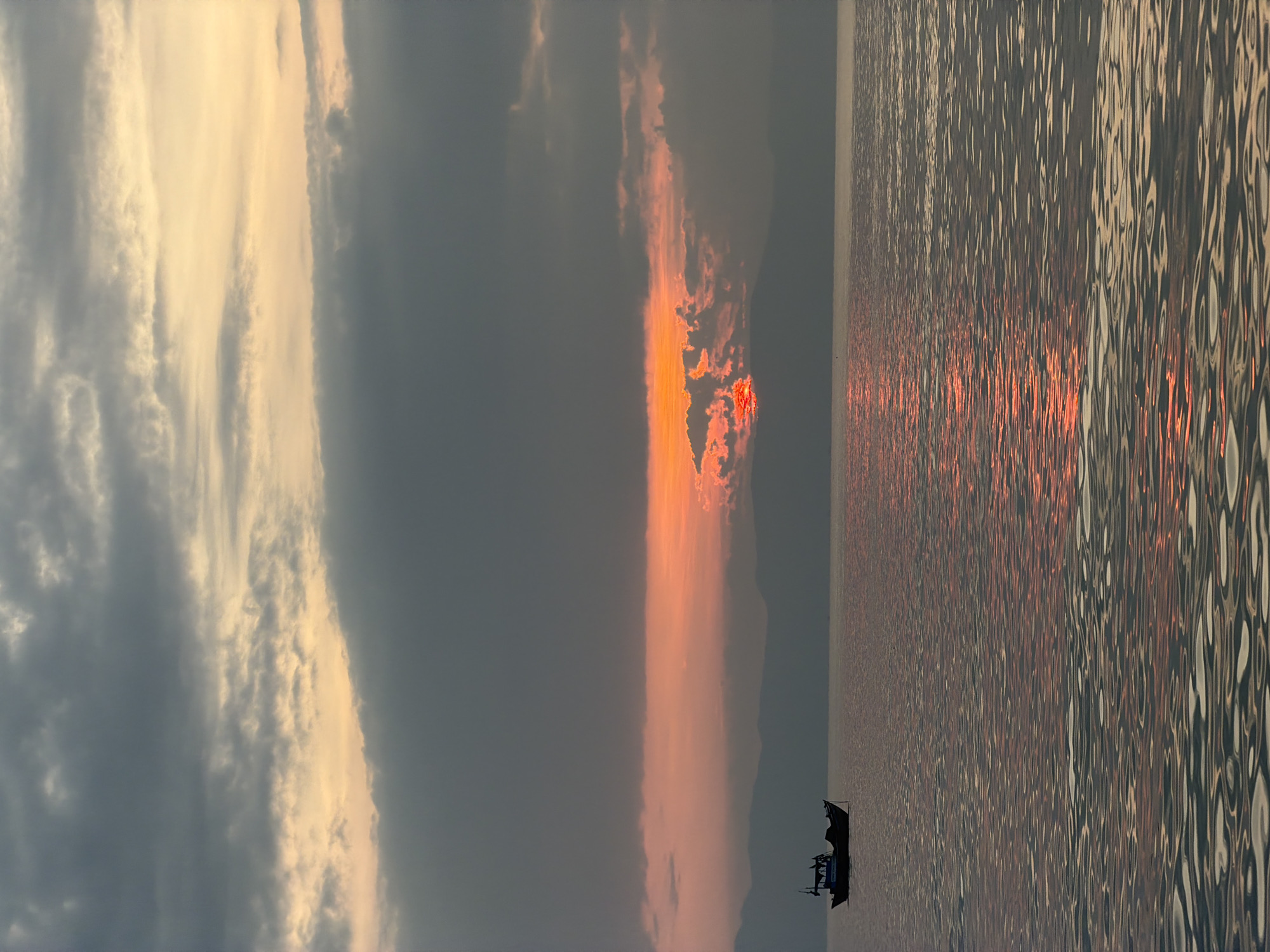 The image depicts a serene seascape at sunset, likely taken from the coast of Ninh Hoa Ward, Khanh Hoa Province, Vietnam. A lone fishing boat, silhouetted against the horizon, floats on calm waters that reflect the vibrant colors of the sky. The sun, partially obscured by dark, heavy clouds, casts an orange and red glow, creating a dramatic contrast with the gray and muted tones of the sky and water. The distant shoreline is marked by a faint mountain range, adding depth to the composition. The overall scene conveys a sense of tranquility and the raw beauty of nature.