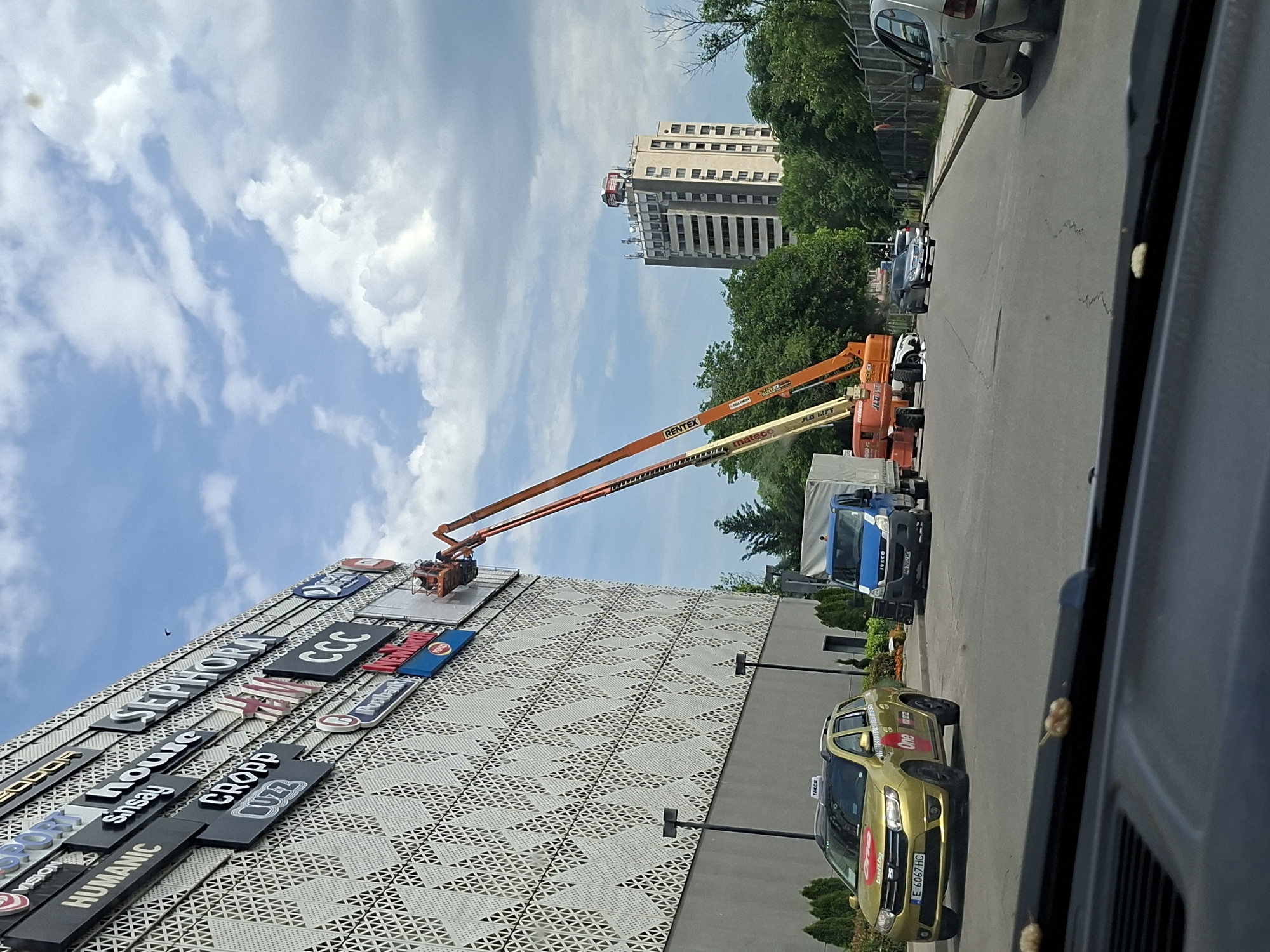 The image, taken in Plovdiv, Bulgaria, depicts a large commercial building, likely a shopping center, undergoing maintenance or installation work on a bright, partly cloudy day. Two extended orange and yellow aerial work platforms, labeled "RENTEX," "mateco," and "JLG LIFT," are positioned against the building's patterned facade. A worker can be seen in the basket of one of the boom lifts, focusing on the various store signs.
The building prominently displays a variety of retail brand signs, including "SEPHORA," "CCC," "Intersport," "HUMANIC," "CROPP," "sinsay," "LIZZ," "sport vision," and "Irobank." On the ground level, a blue "IVECO" truck is parked, and a gold/yellow Dacia Logan taxi, branded "One" and "taxit.bg," is visible on the adjacent road, displaying the Bulgarian license plate "E 6067 HC." Other vehicles are parked further down the road. In the background, beyond a line of green trees, a tall, light-colored building with a visible "BILLA" sign on its roof stands against the sky. The scene suggests a busy commercial area during daytime hours.